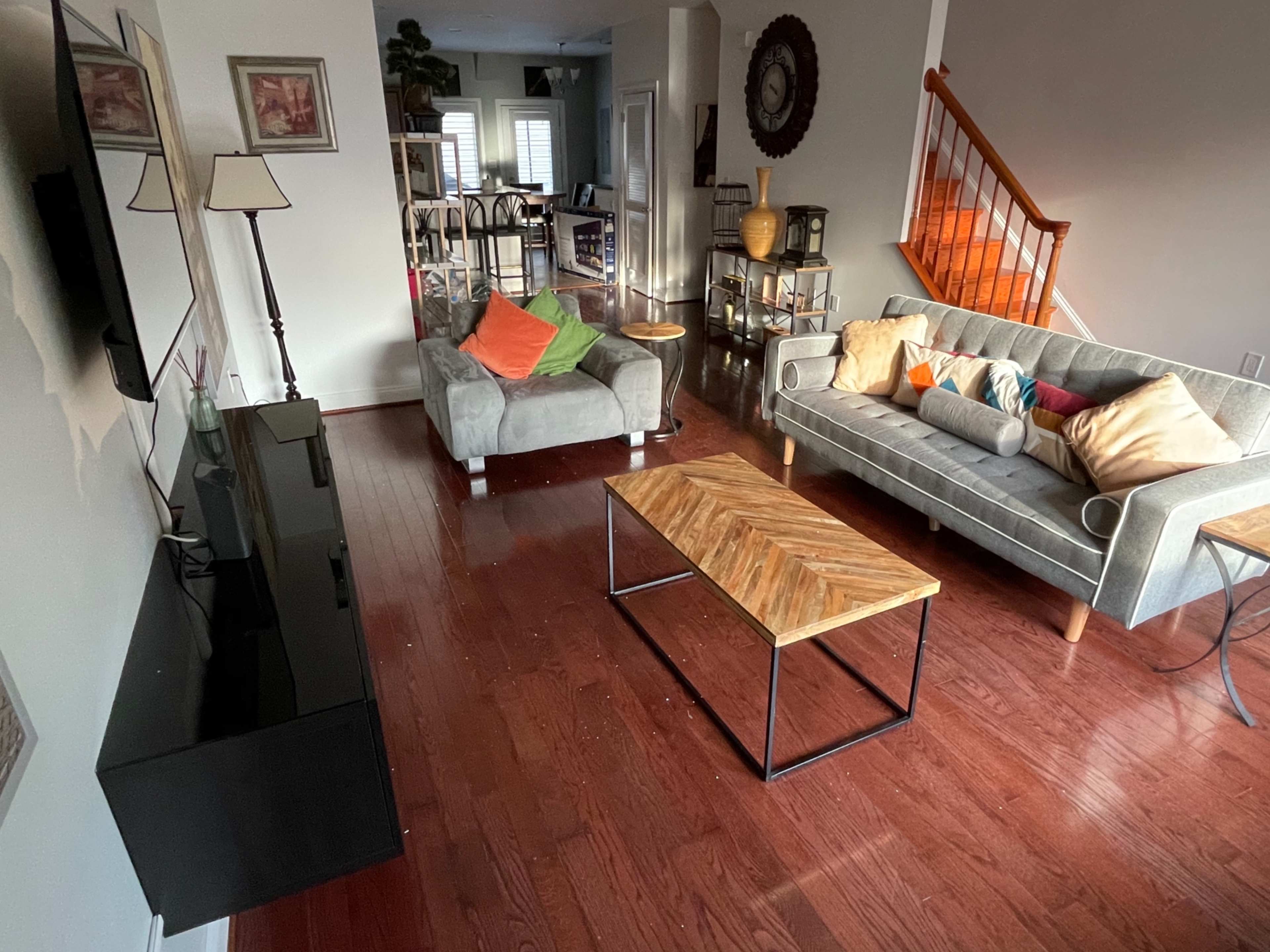 The living room features a gray sectional sofa and a light gray couch with colorful pillows, a wooden coffee table, a television on the wall, and hardwood flooring.
