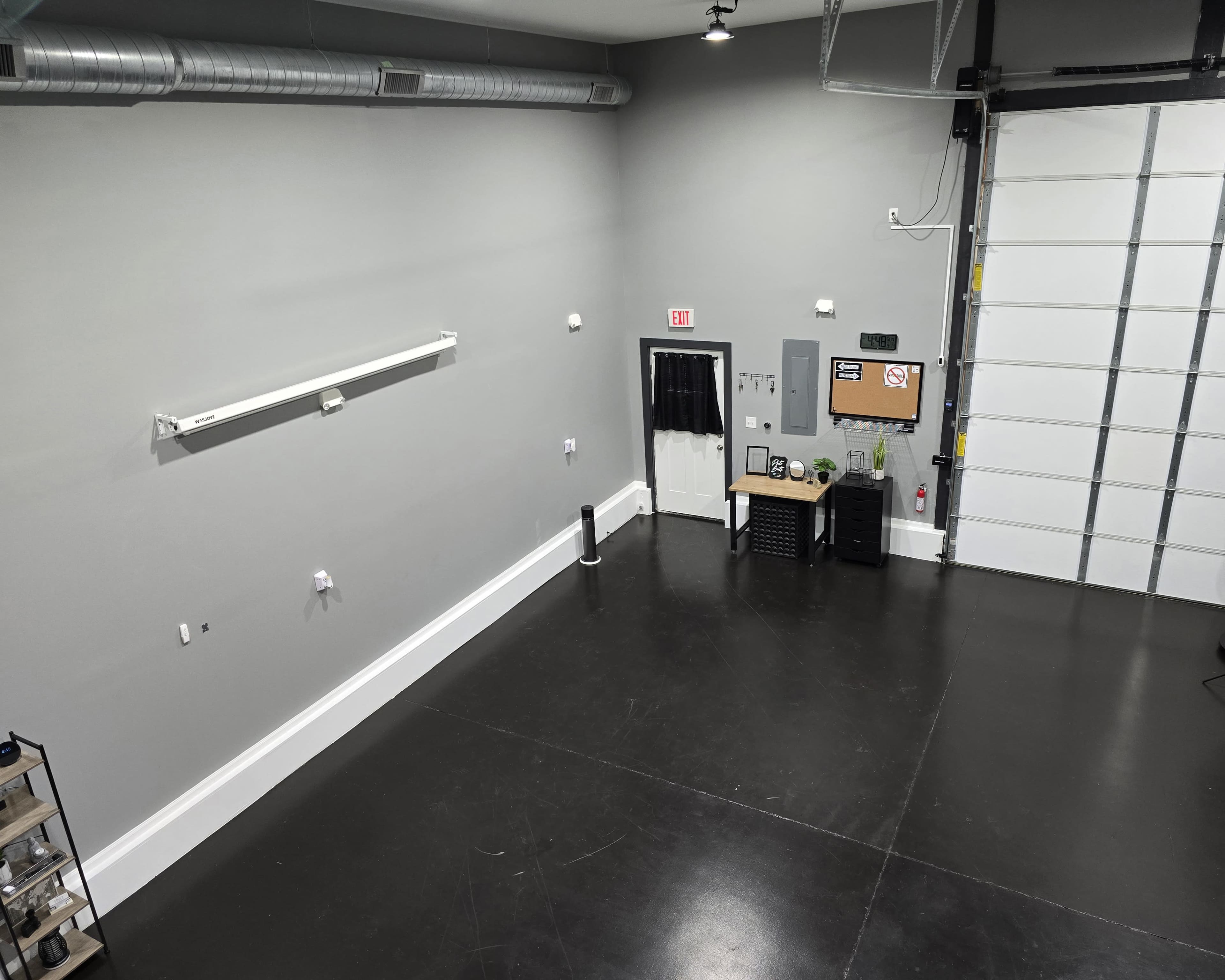 The image shows an empty, spacious garage with gray walls, a large white garage door, and a minimalistic setup featuring a small table and storage unit.