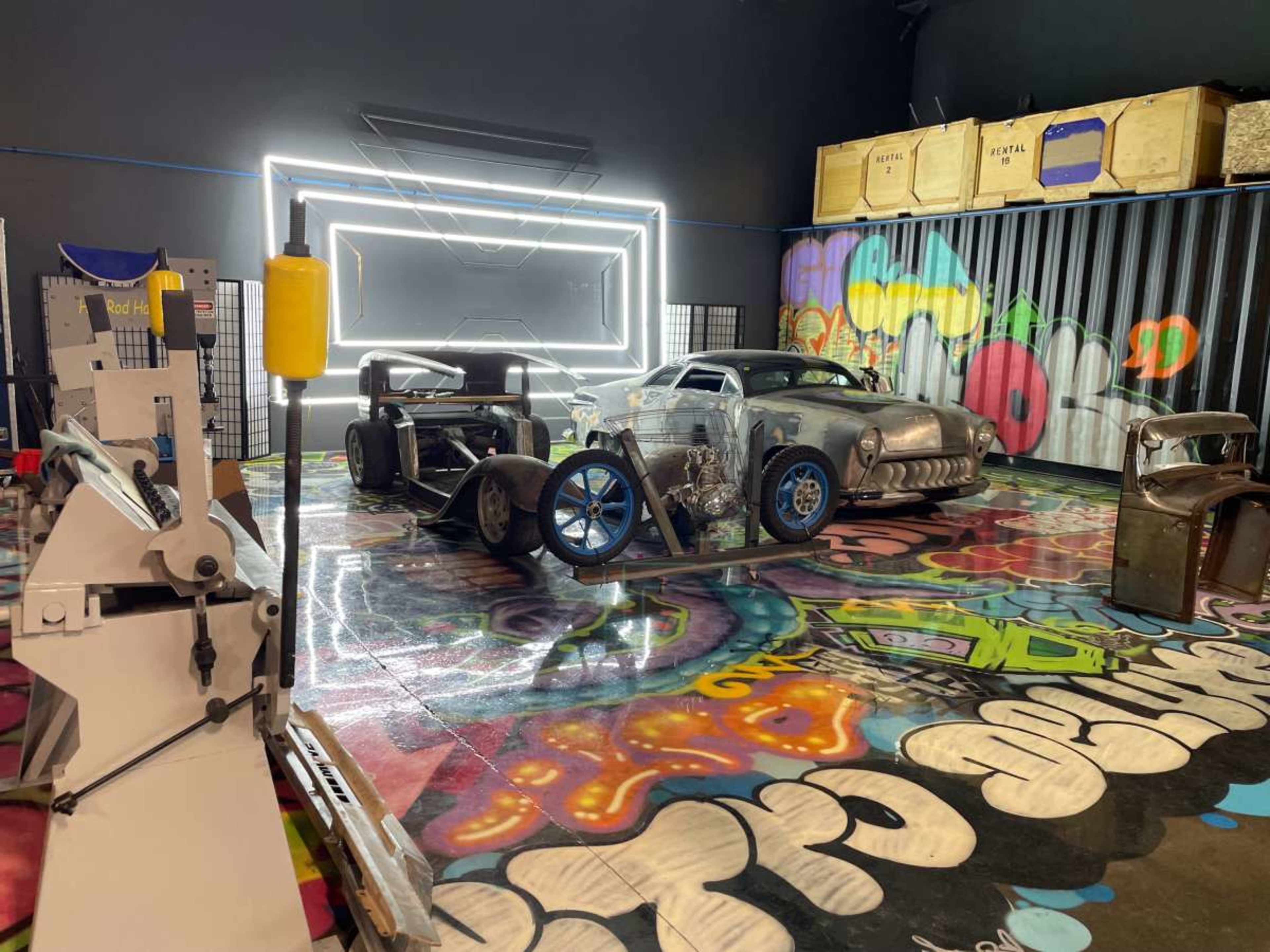 The image shows a colorful workshop space featuring several vintage cars, graffiti art on the floor, and various tools and equipment in the foreground.