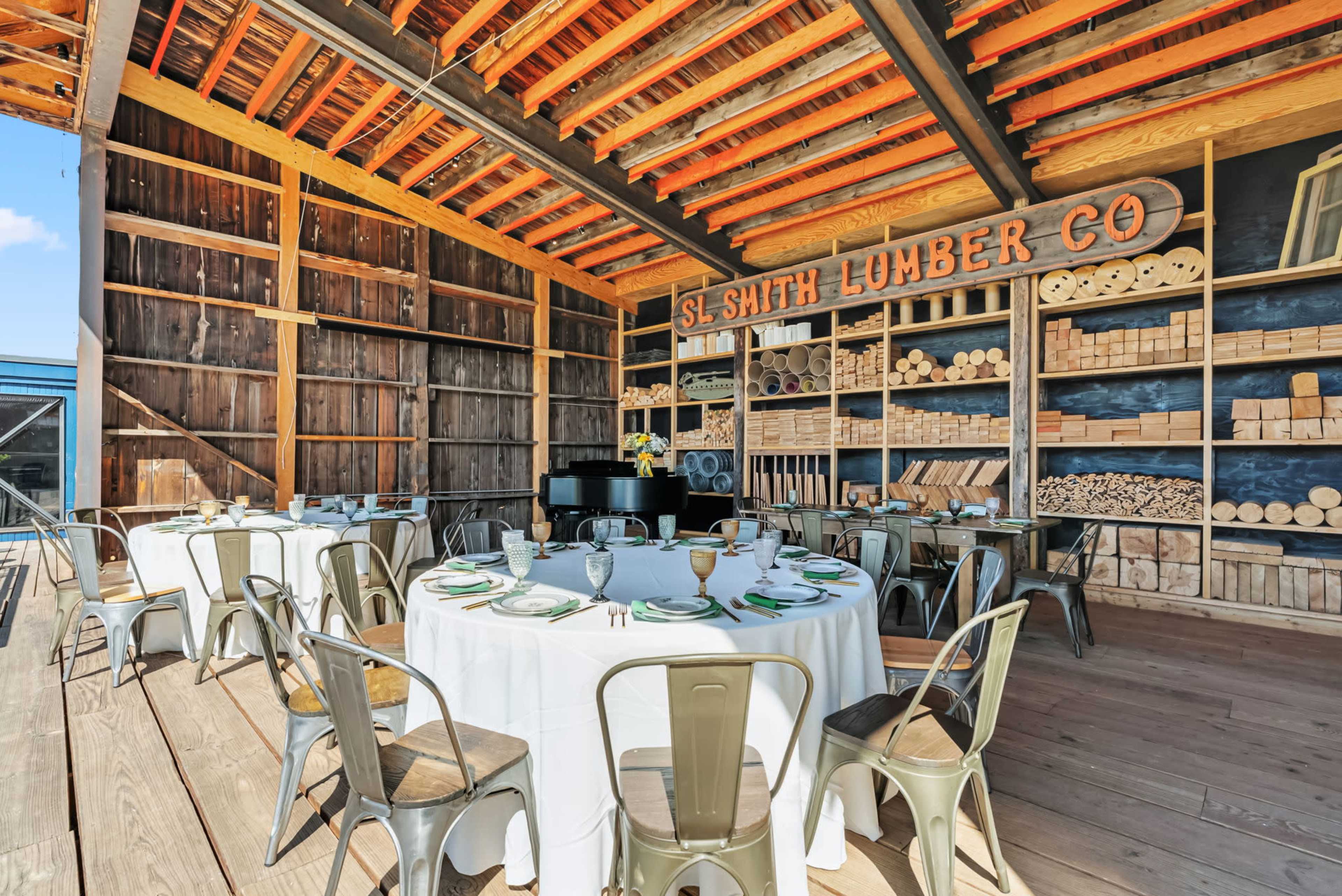 The image shows a rustic barn interior set up for a dining event, featuring round tables with white linens and metal chairs, alongside shelves stocked with lumber and supplies.