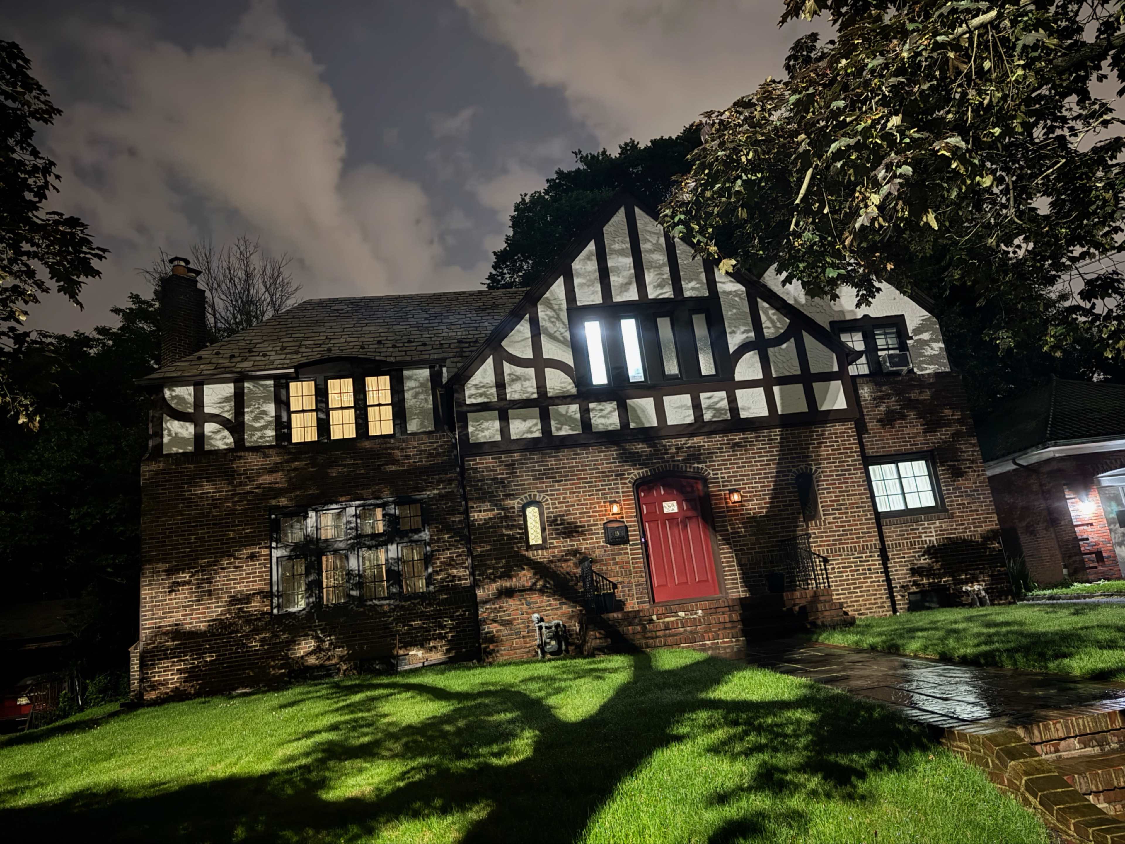 A brick house with a Tudor-style design features a red door and illuminated windows, set against a night sky with clouds.
