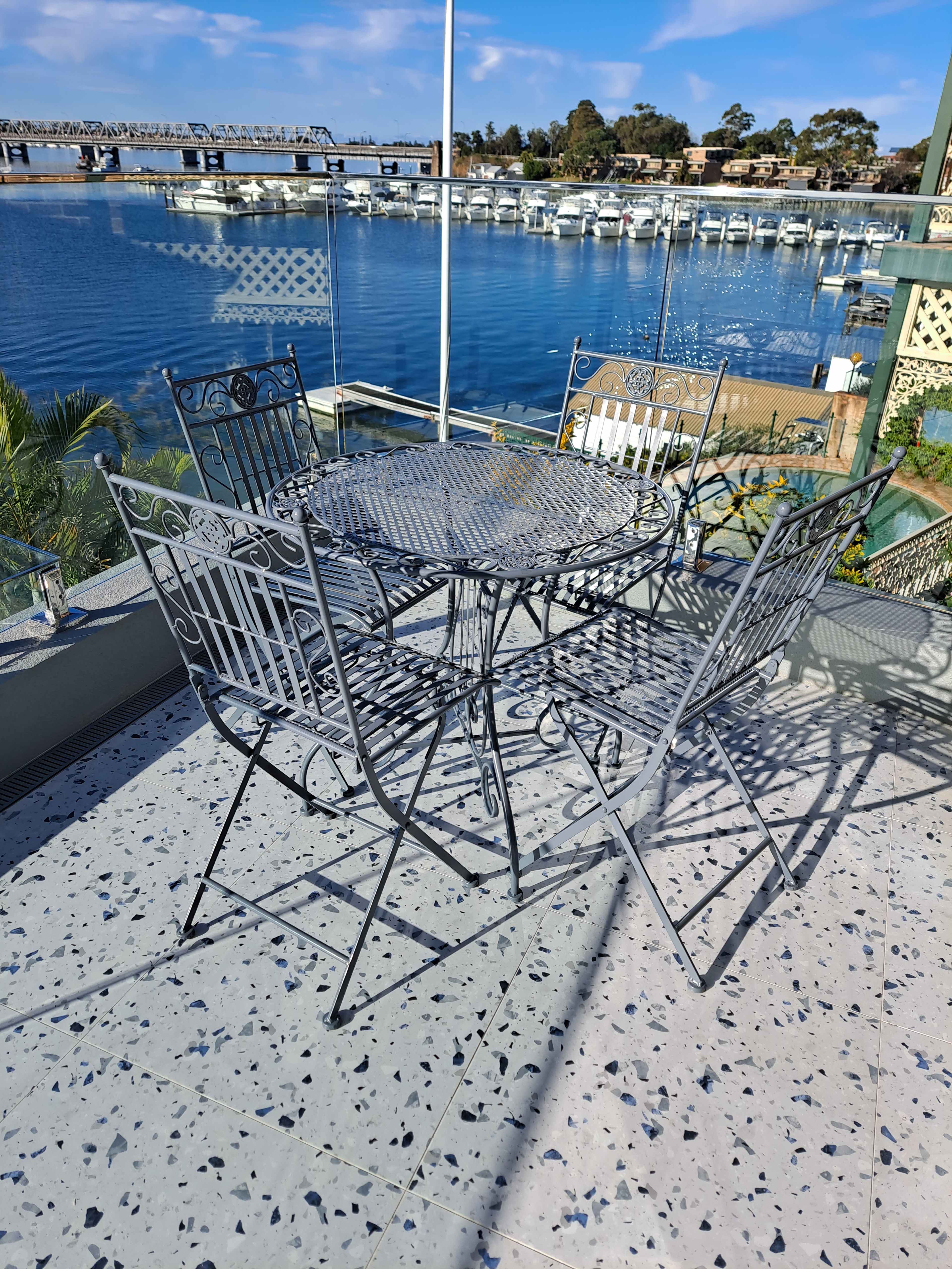 A round metal table with four matching chairs is positioned on a terrazzo floor, overlooking a marina with moored boats and a clear blue sky.