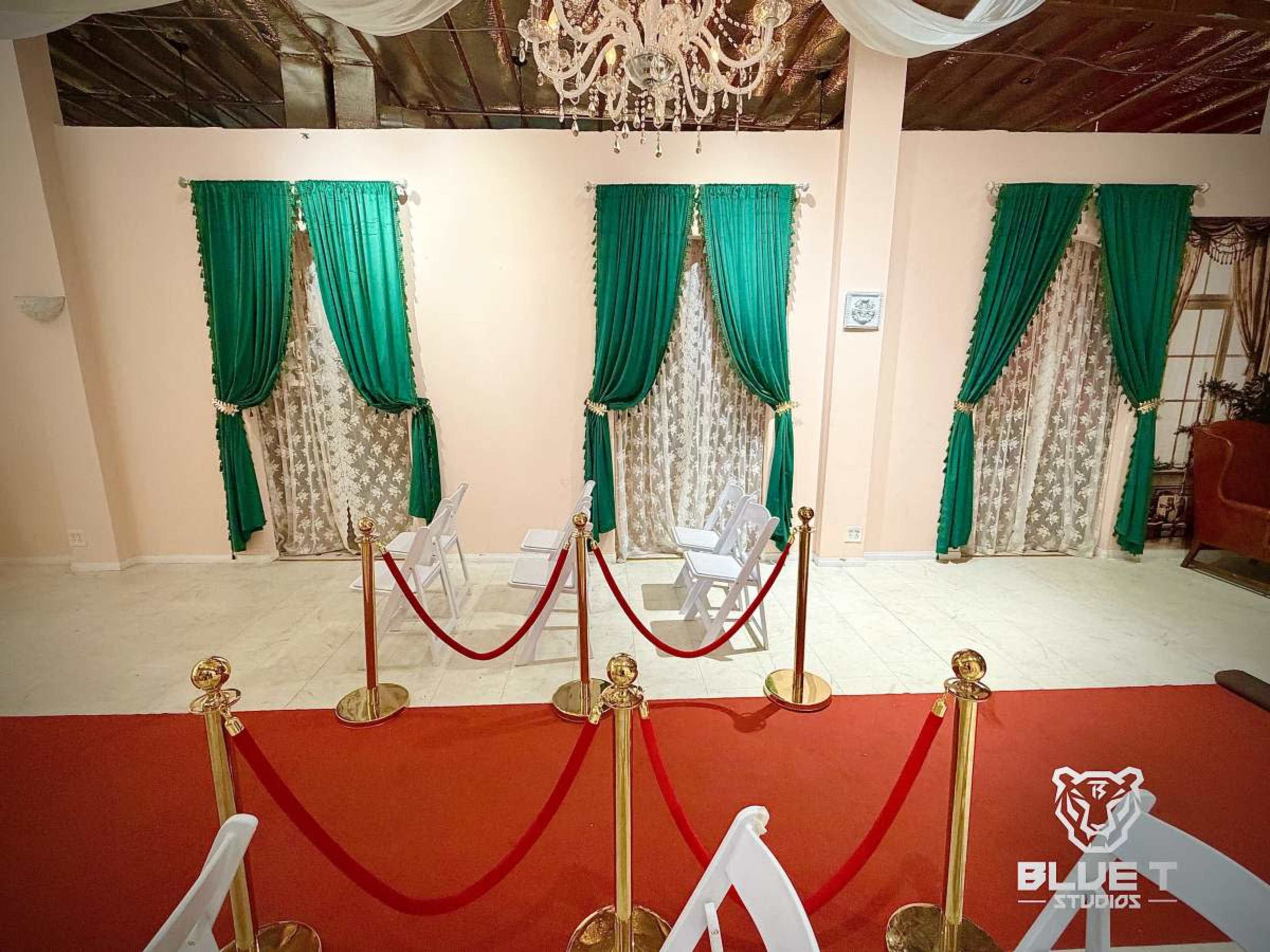 The image shows a room with a red carpet leading to two windows adorned with green curtains, framed by sheer drapes, and a stanchion barrier in front.