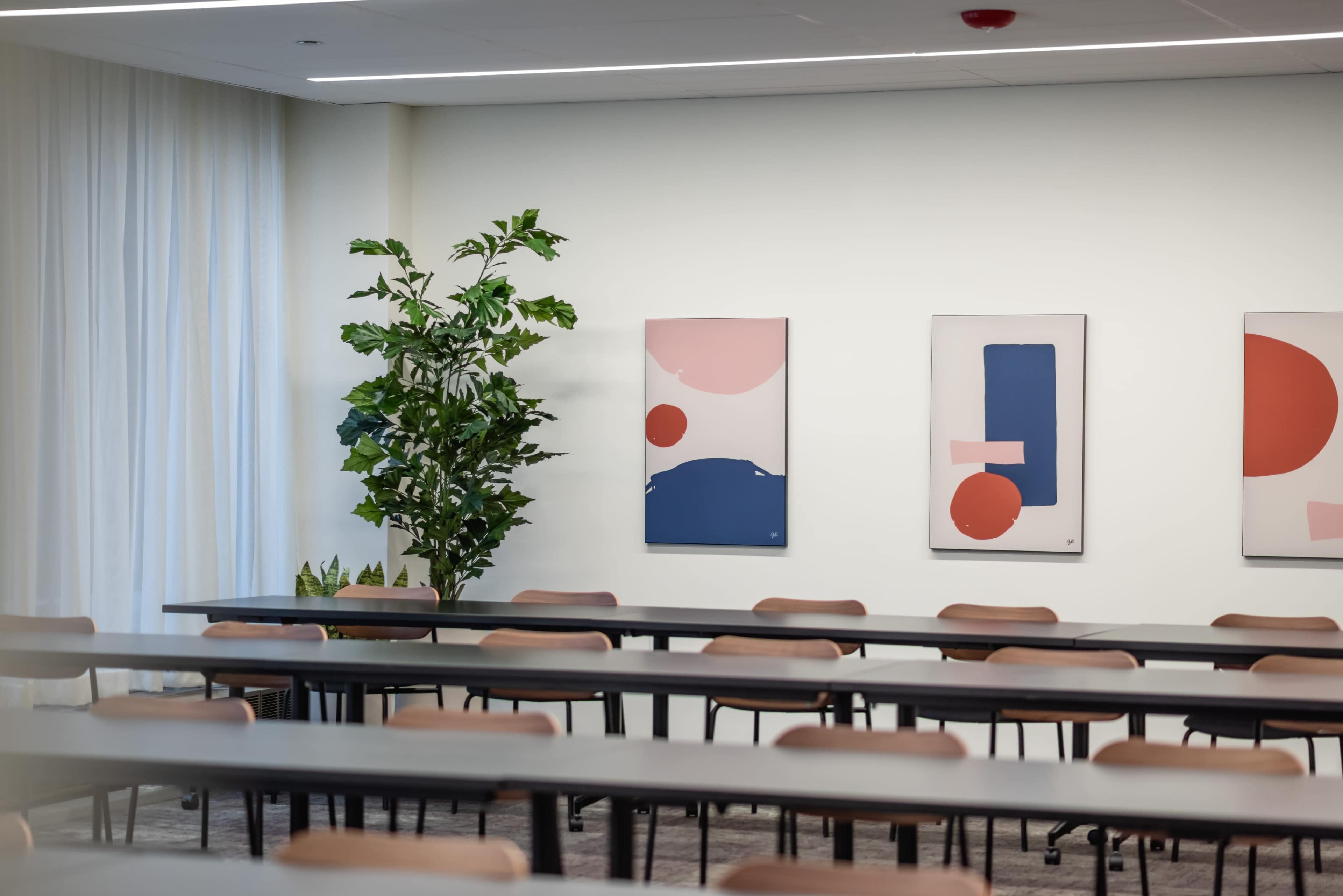 A conference room features rows of tables and chairs, along with abstract wall art and a potted plant in the corner.