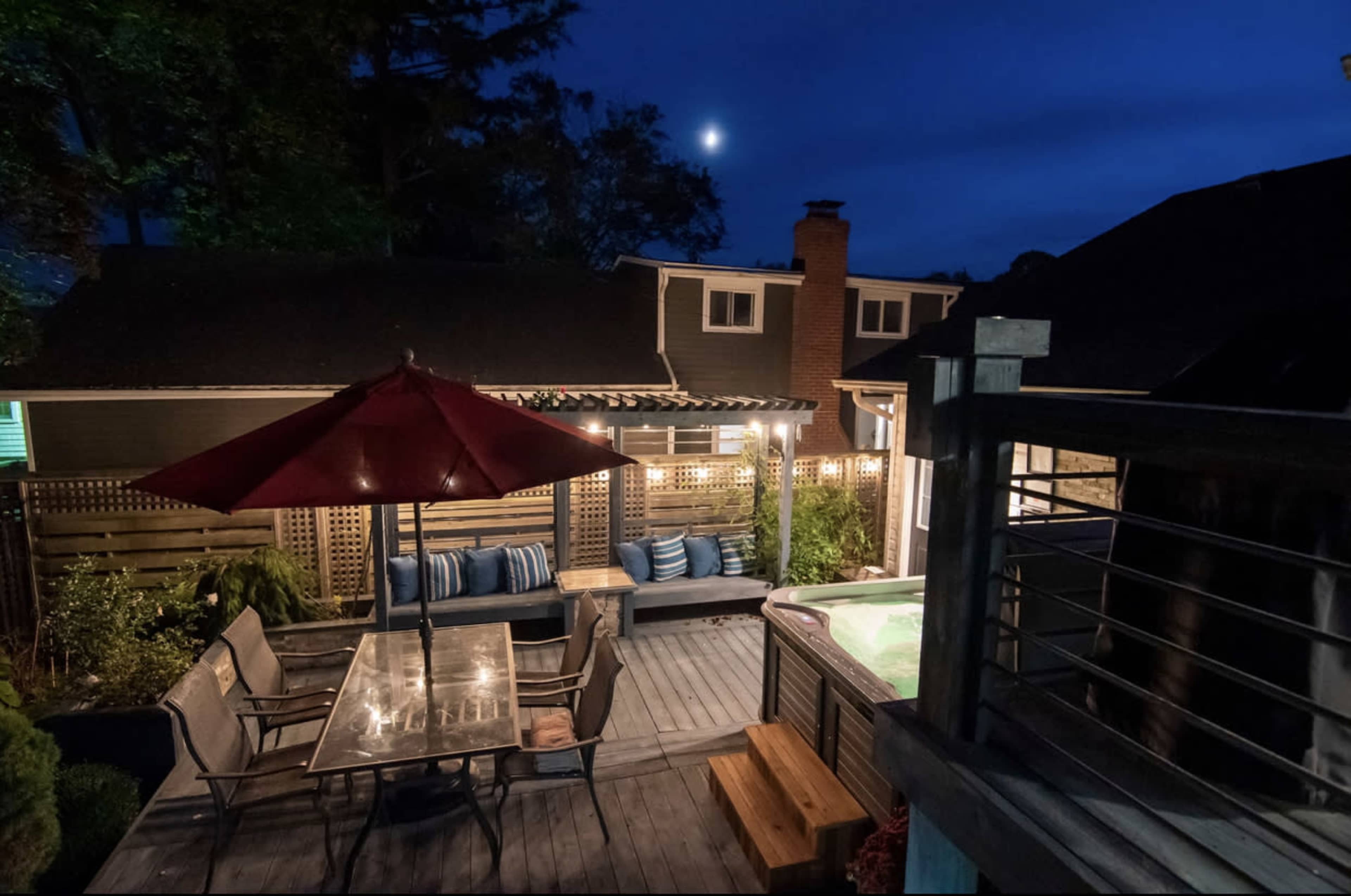 A backyard deck features a dining table under an umbrella, a hot tub, and string lights illuminated at night with the moon visible in the sky.