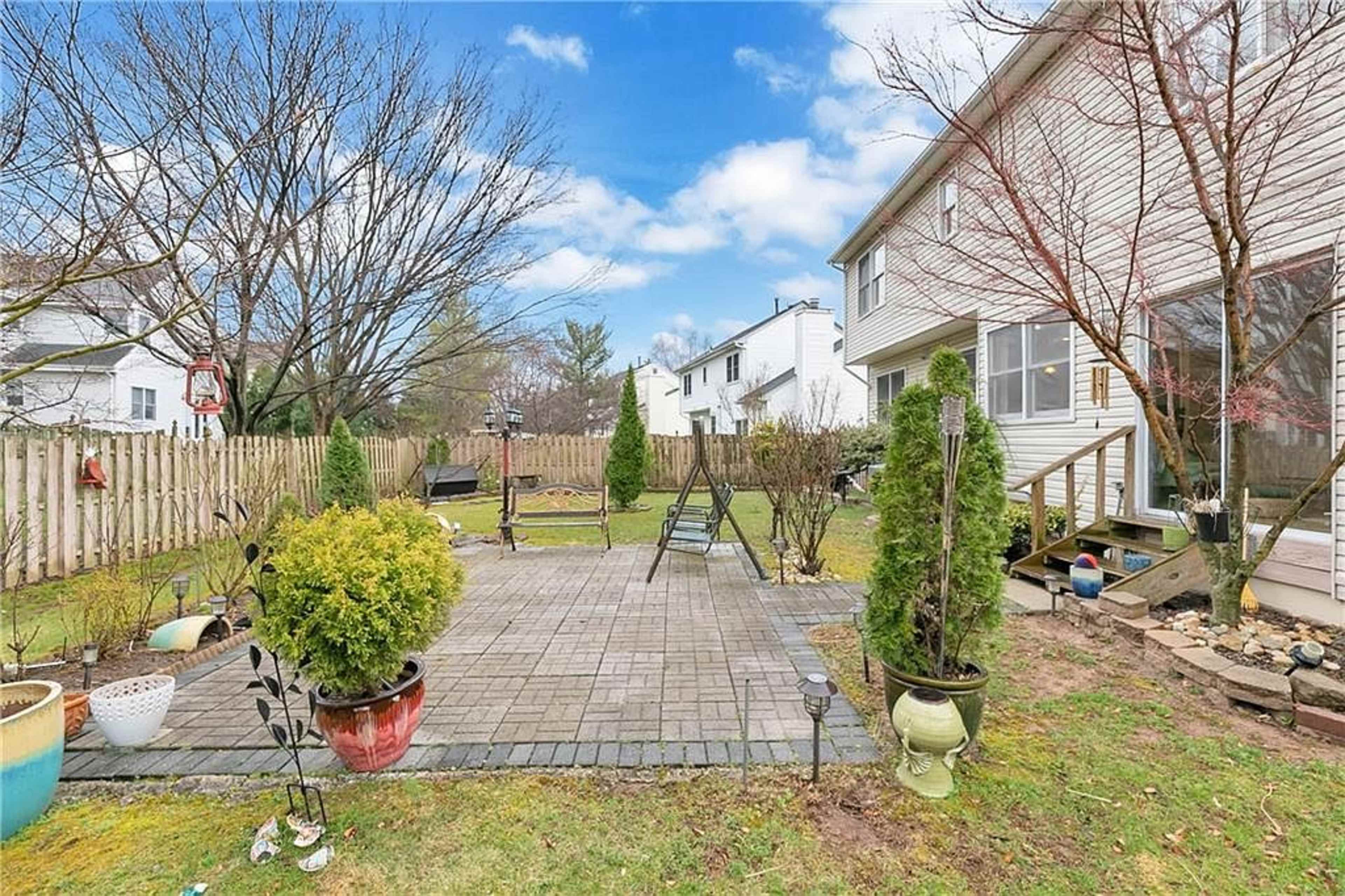The image shows a backyard with a stone patio, several potted plants, and a wooden deck leading to a house, surrounded by a wooden fence and grass.