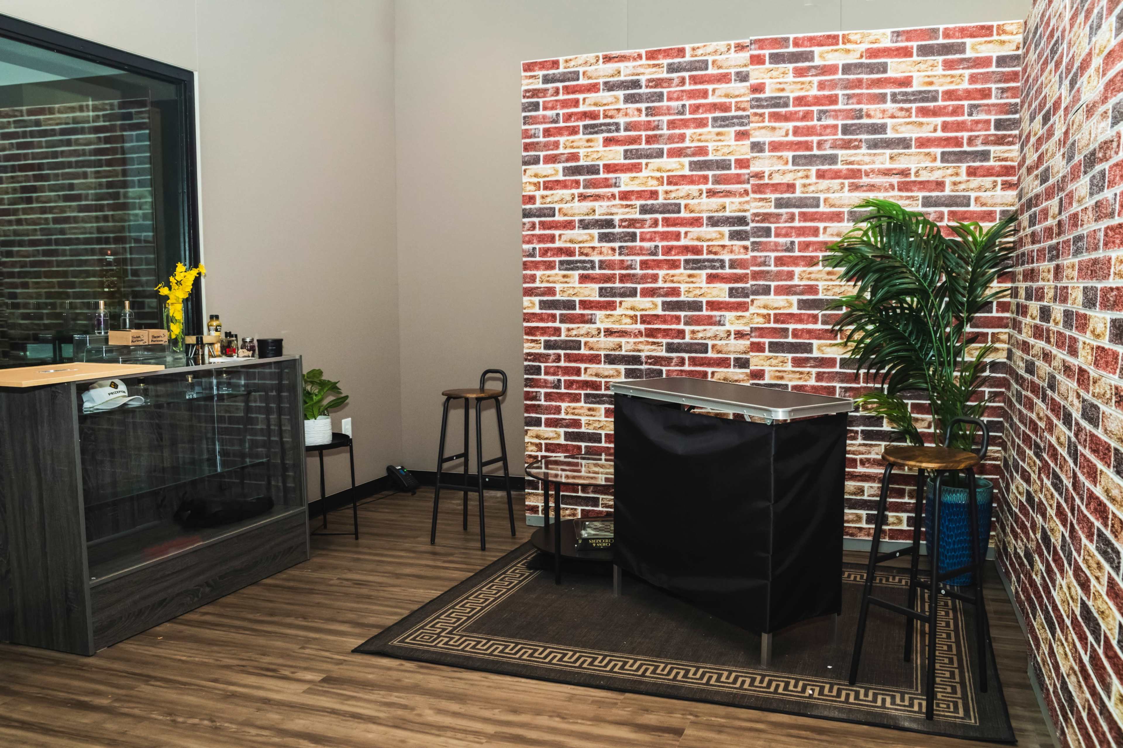 A small room with a brick-patterned wall, a glass cabinet, two stools, a black cart, and a potted plant.