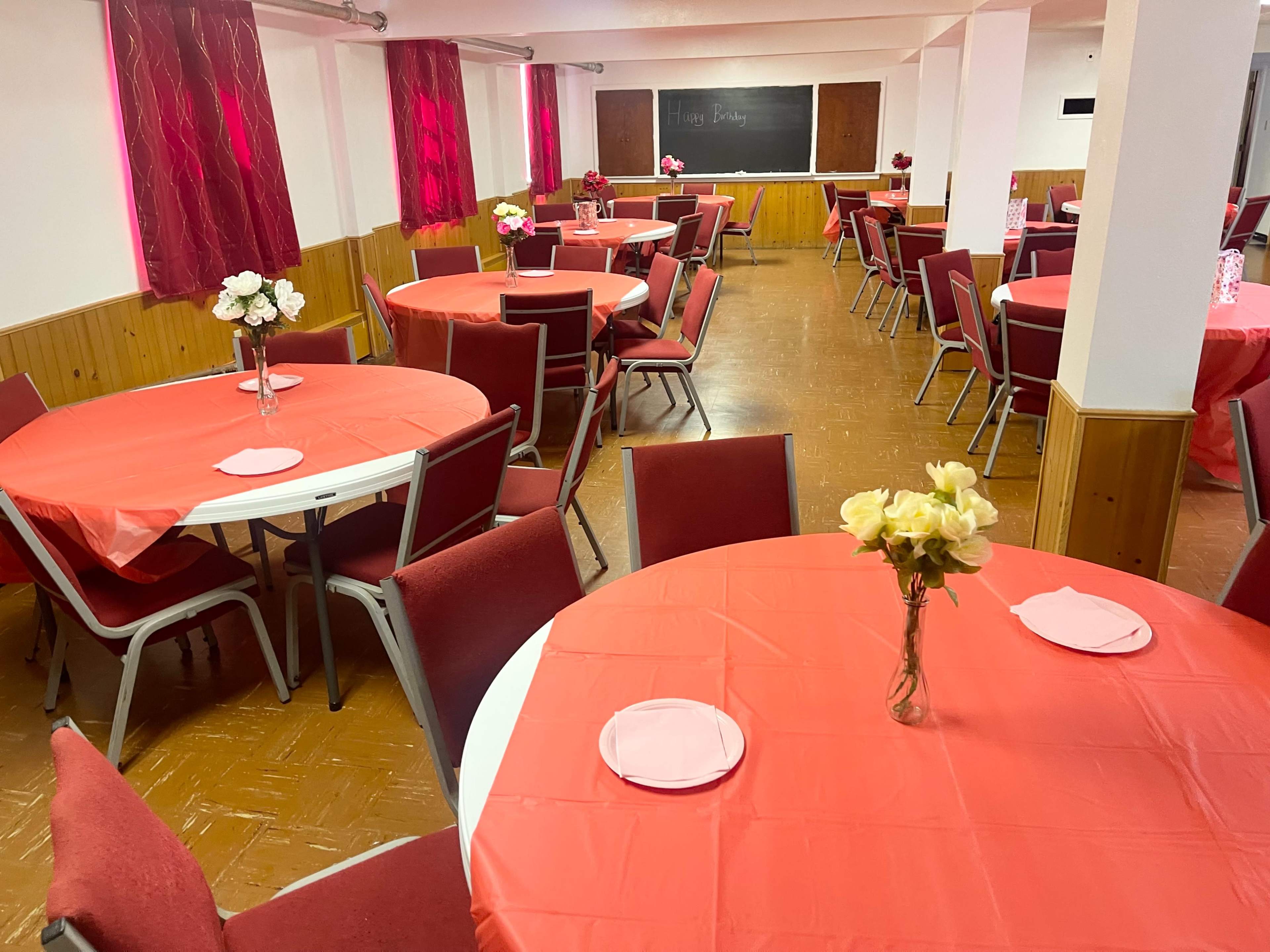 The image shows a banquet room decorated with red tablecloths and centerpieces of flowers on round tables, arranged for an event.