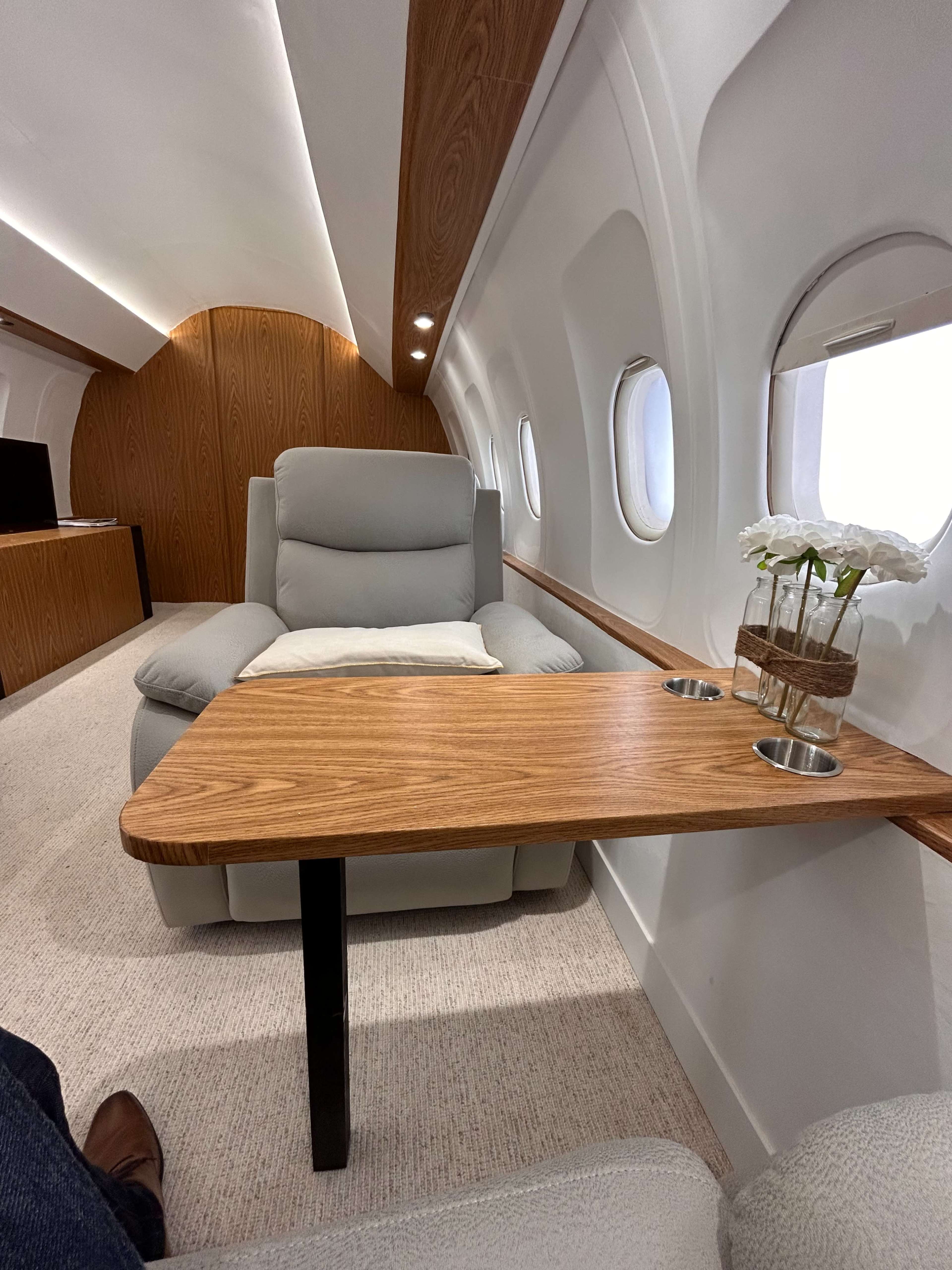 A luxurious airplane interior with a gray armchair, a wooden table with cup holders, and large windows.