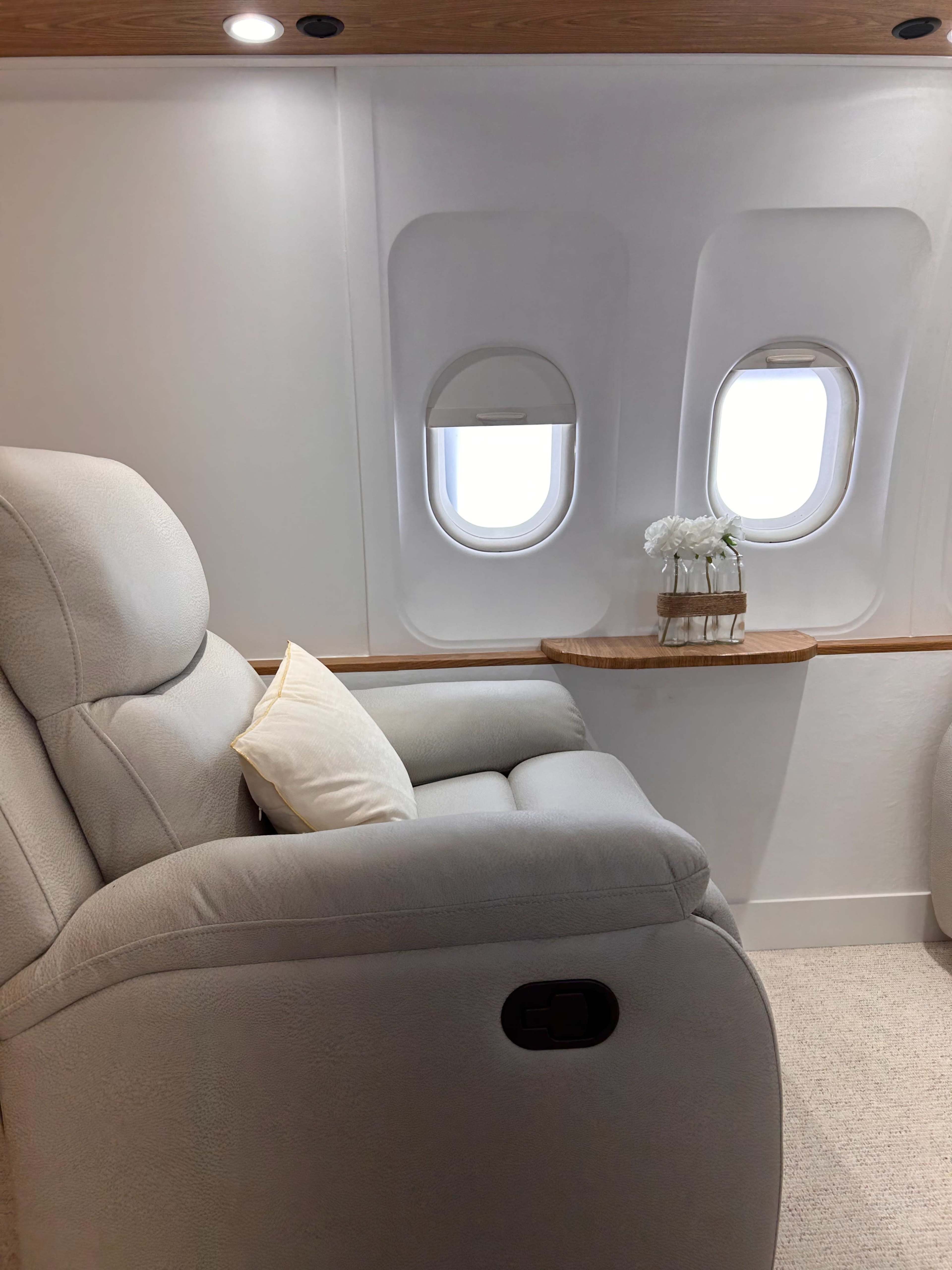 A recliner sits next to two airplane windows, with a small table holding a vase of flowers in the background.