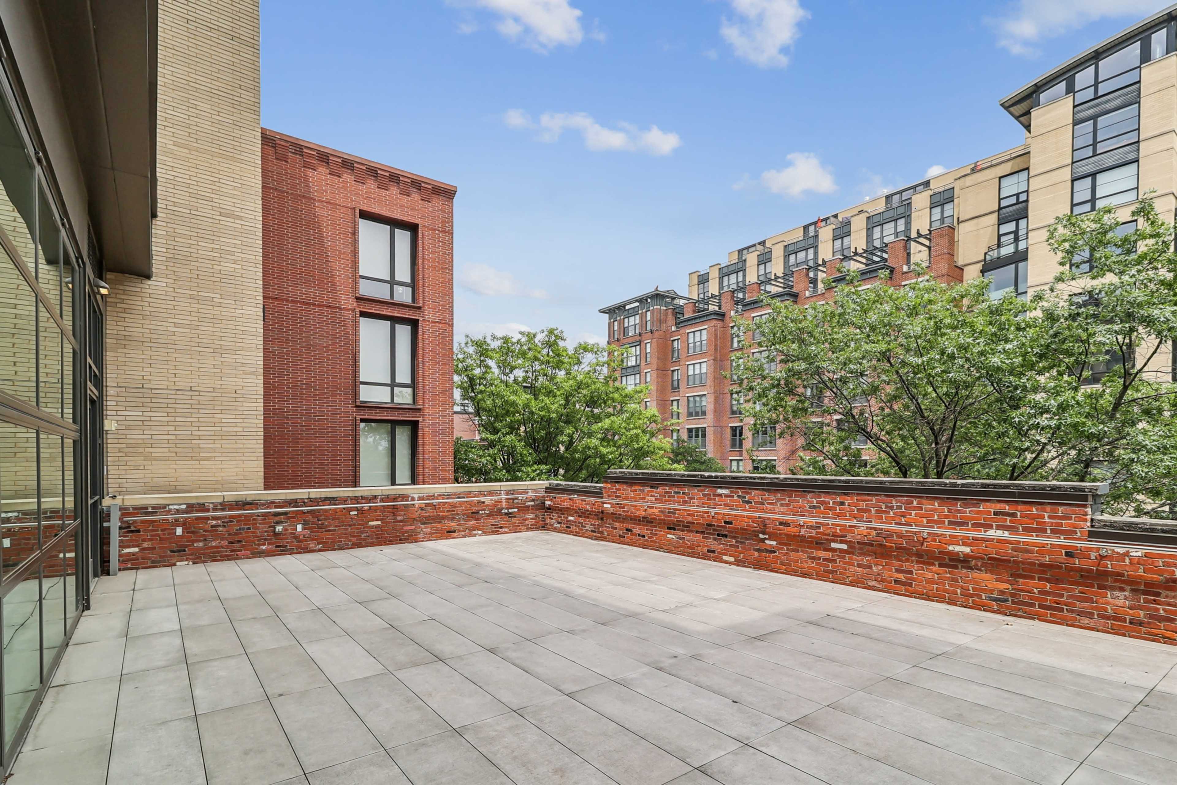 The image shows a spacious brick terrace with tiled flooring, surrounded by trees and apartment buildings.