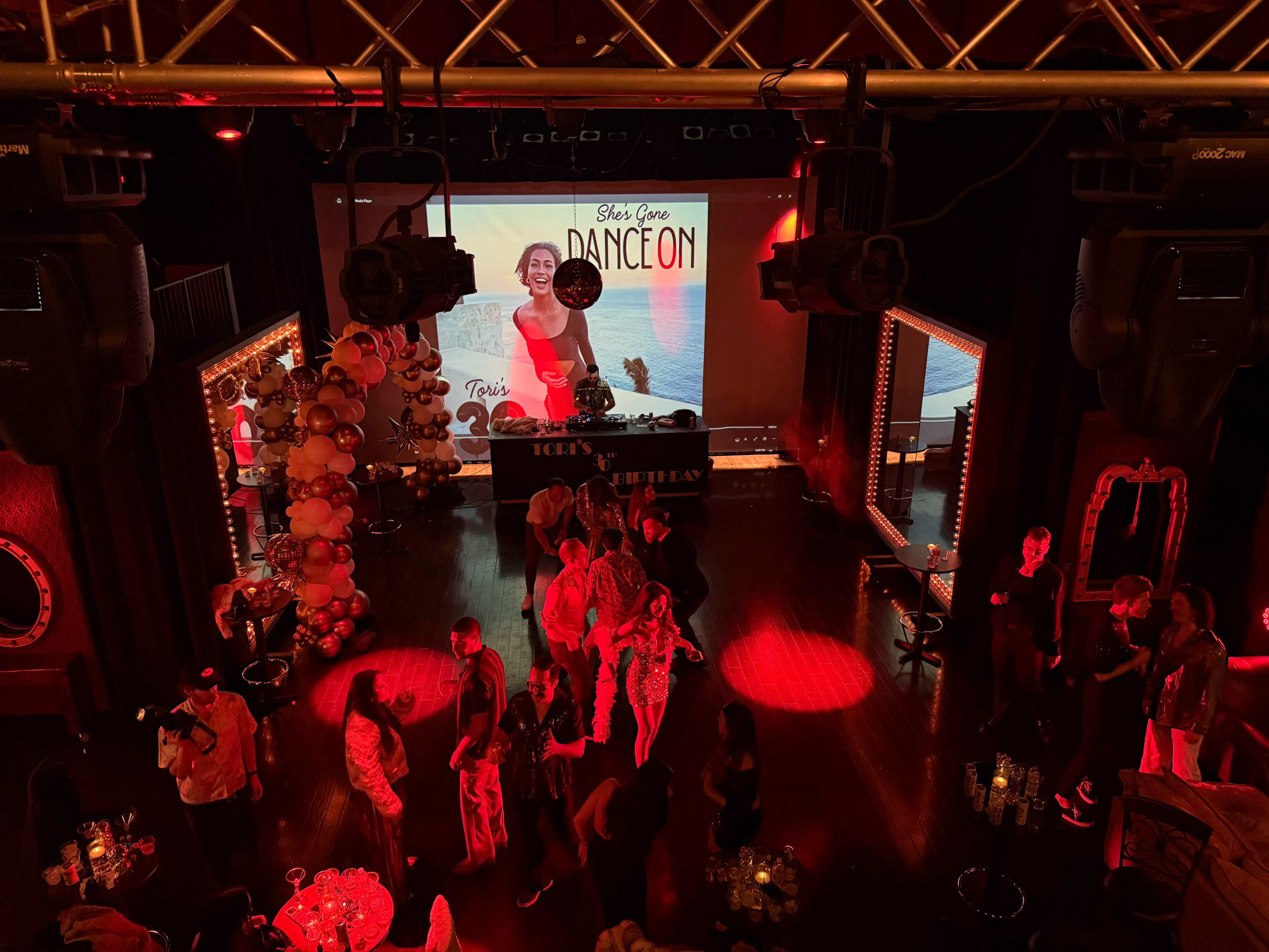 The image shows a lively indoor event space with guests mingling under red lighting, a large screen displaying a promotional image, and decorative elements such as balloons.