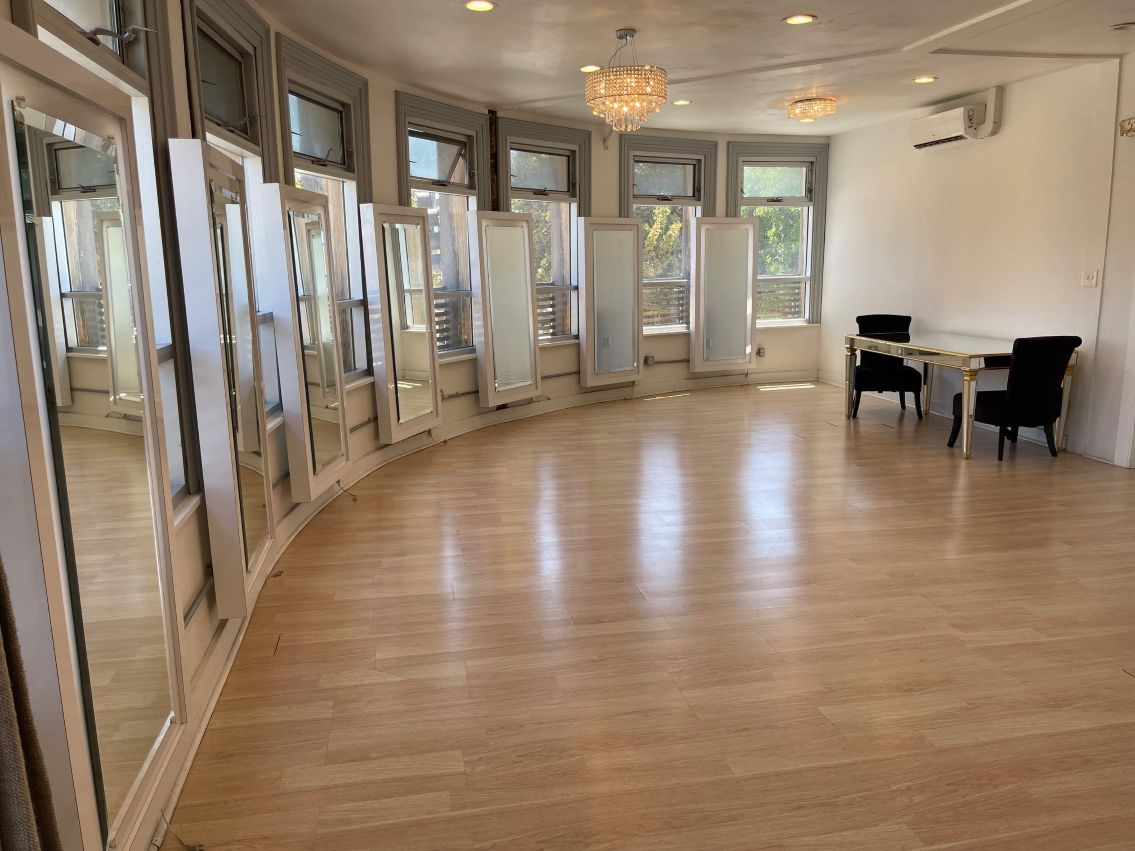 The image shows a spacious room with a curved wall of mirrors, hardwood flooring, and a table with two chairs near the windows.