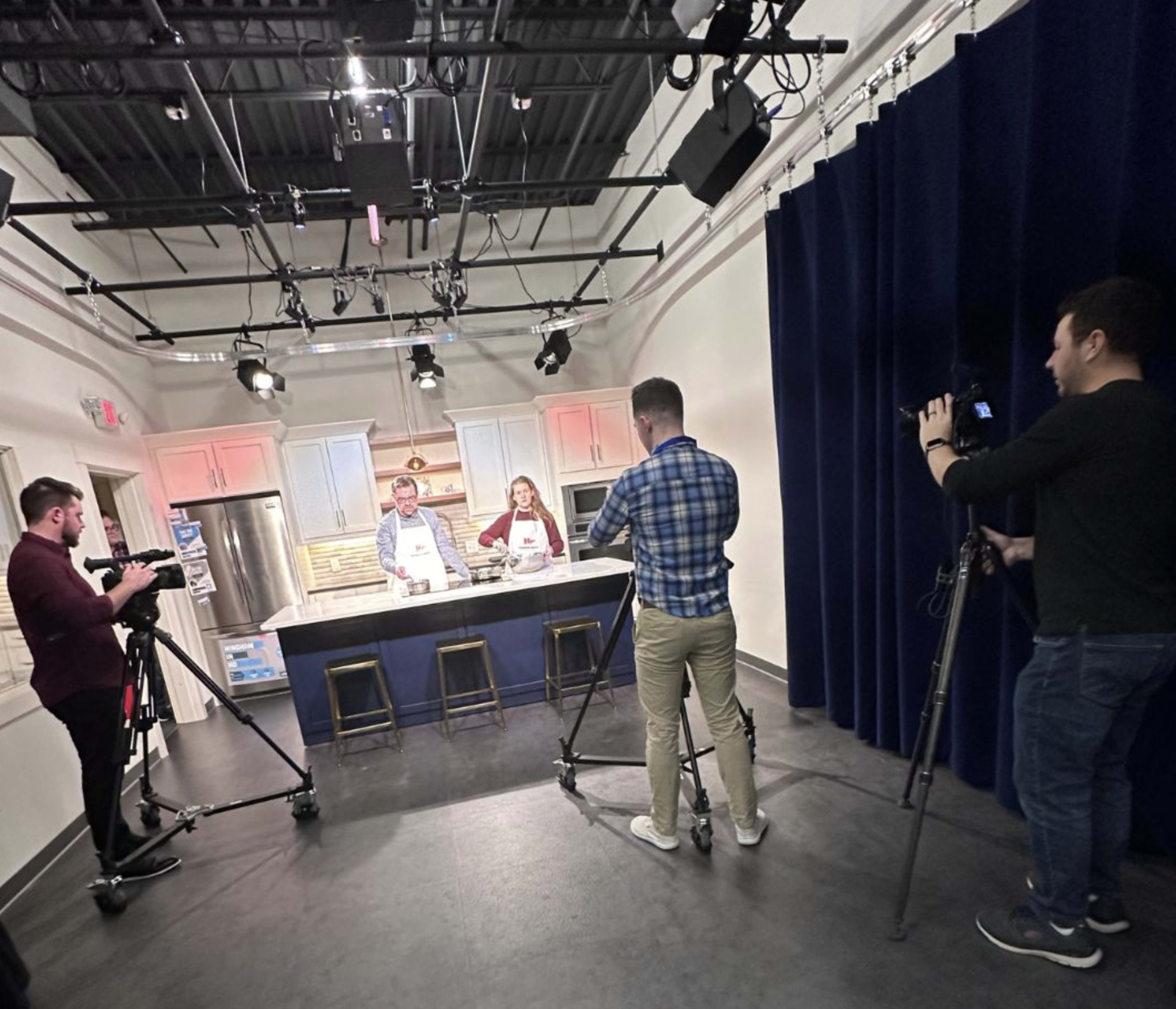 Two people are cooking at a kitchen set while three others are filming them with cameras.