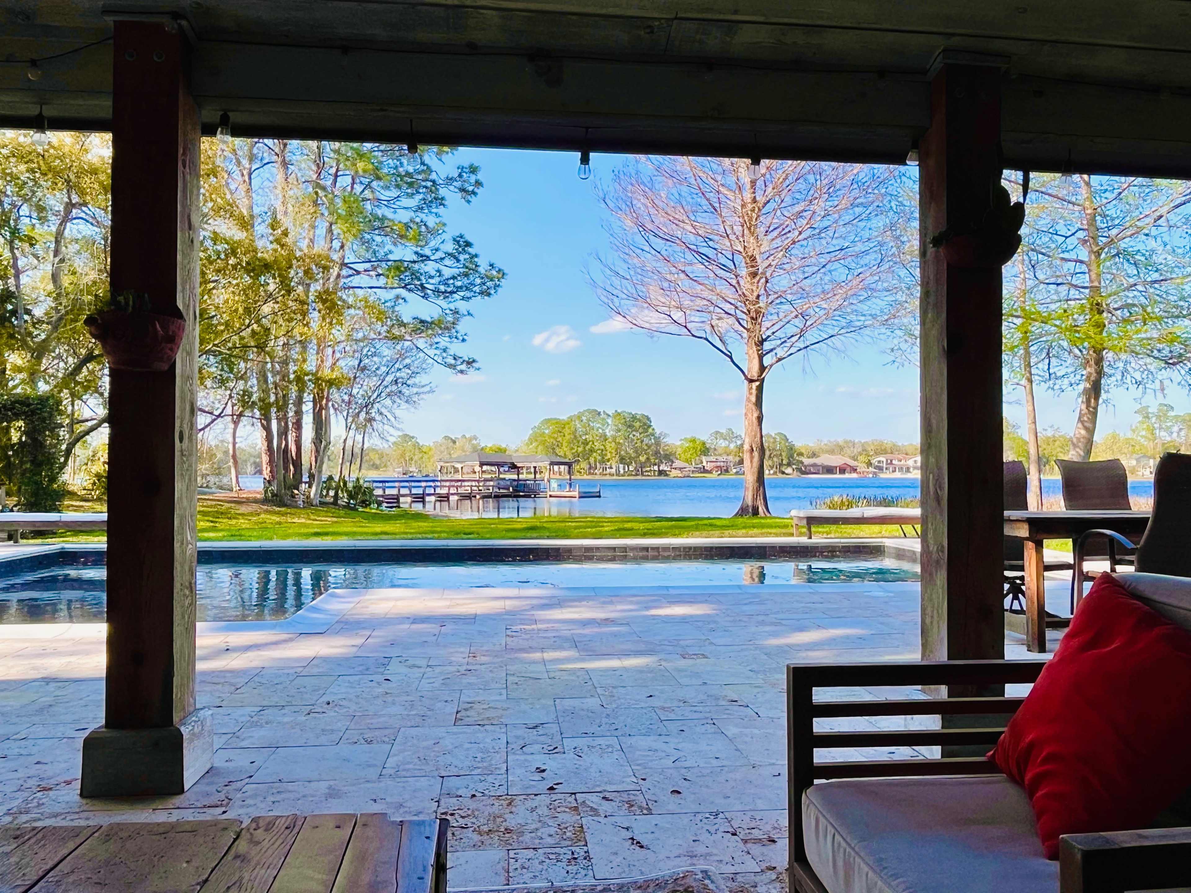 A view from a covered porch overlooking a pool and lake surrounded by trees.