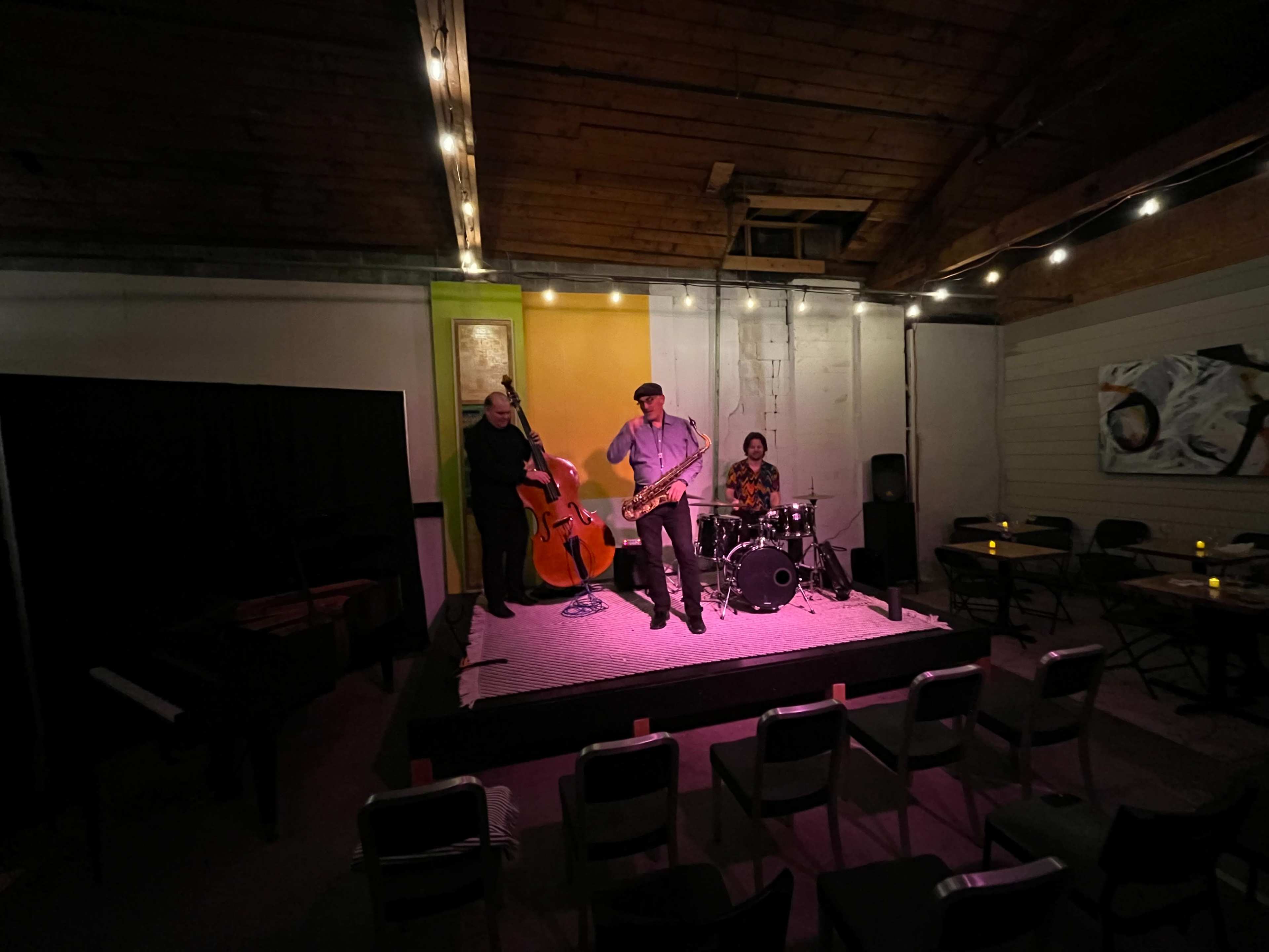 A jazz trio performs on a small stage in a dimly lit room, featuring a saxophonist, a double bassist, and a drummer.