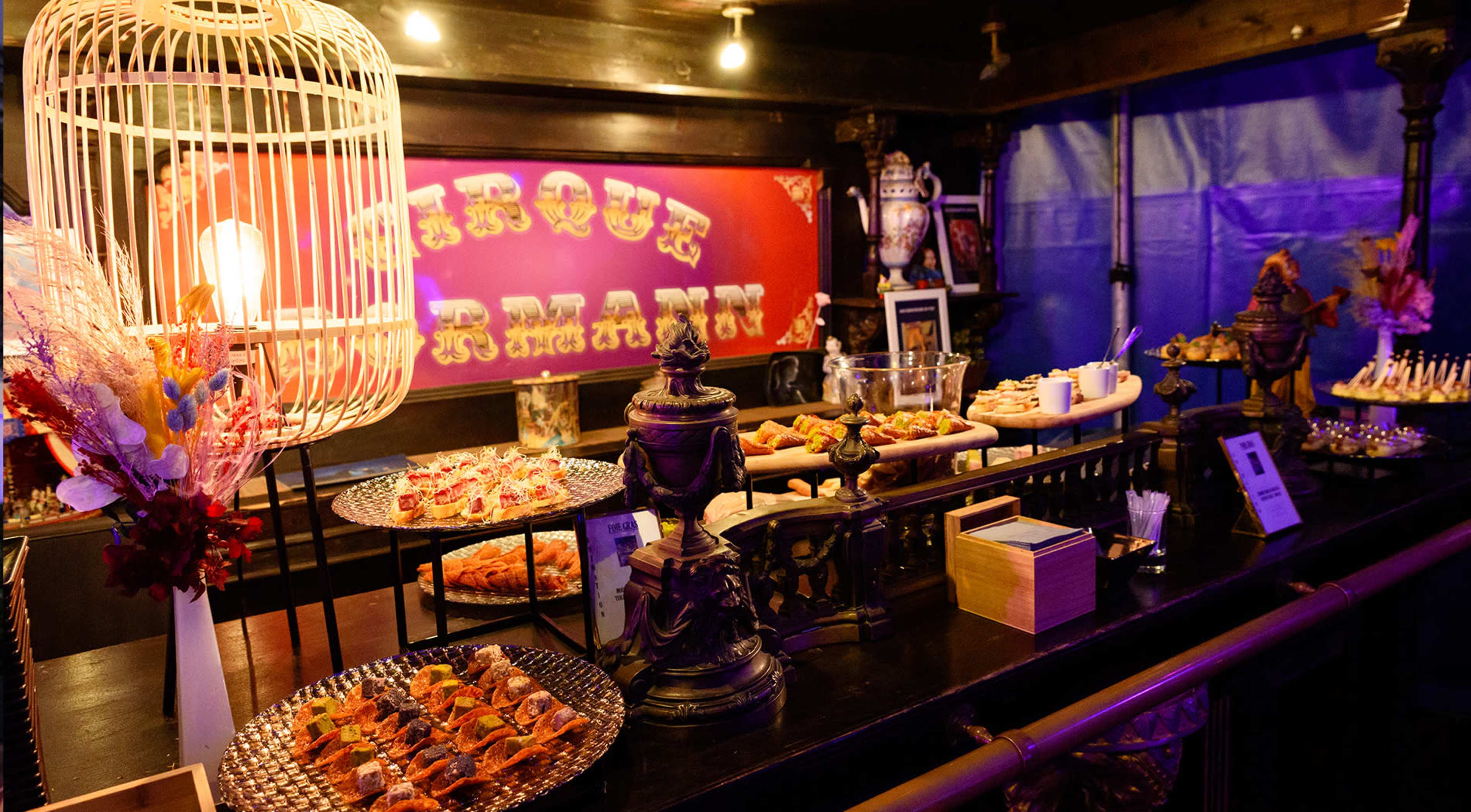 A buffet table displays a variety of appetizers, desserts, and decorative elements under warm lighting in a themed setting.
