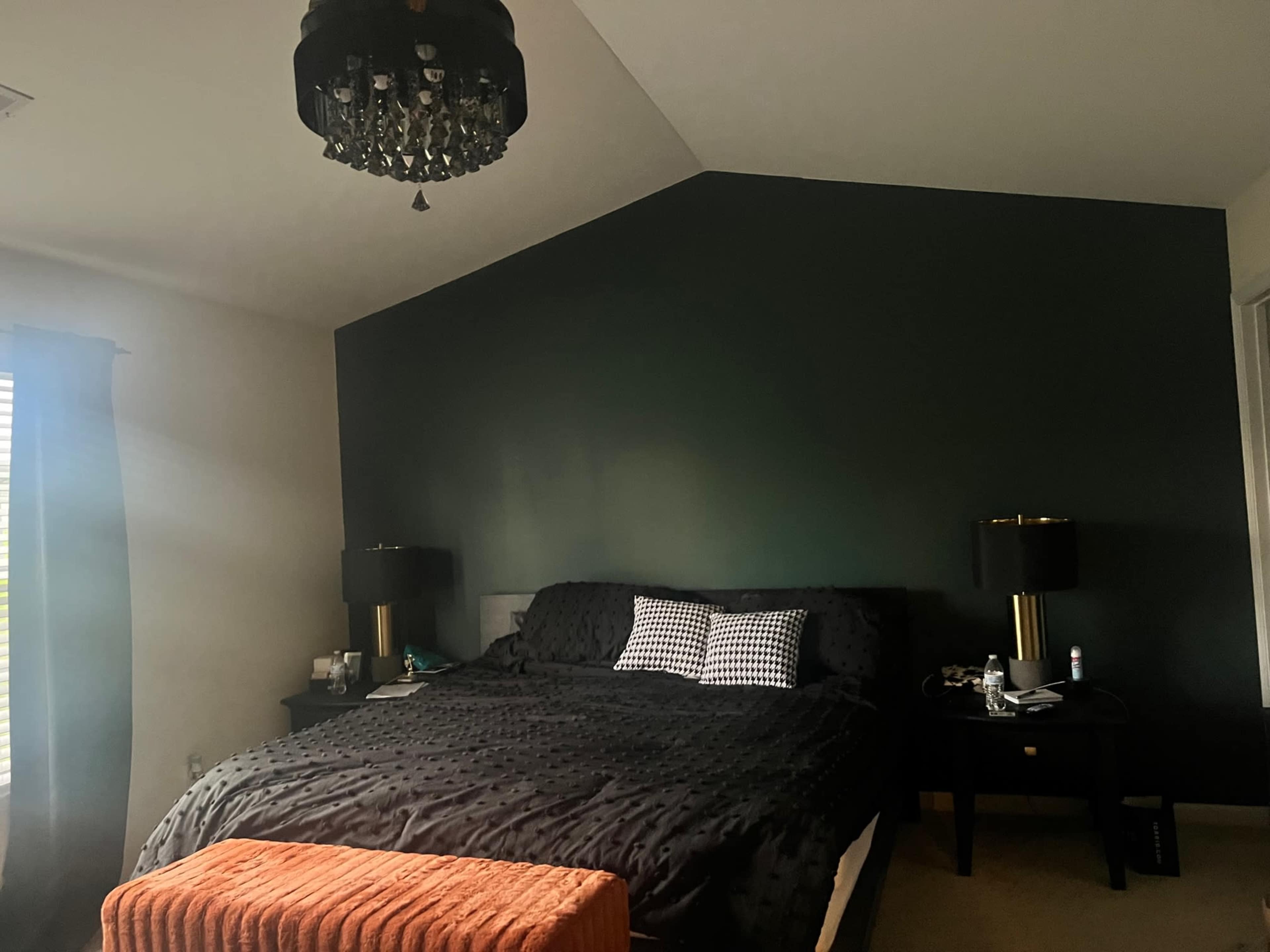 A dark-colored bedroom features a bed with black linens, two bedside tables with lamps, and an orange ottoman at the foot of the bed.