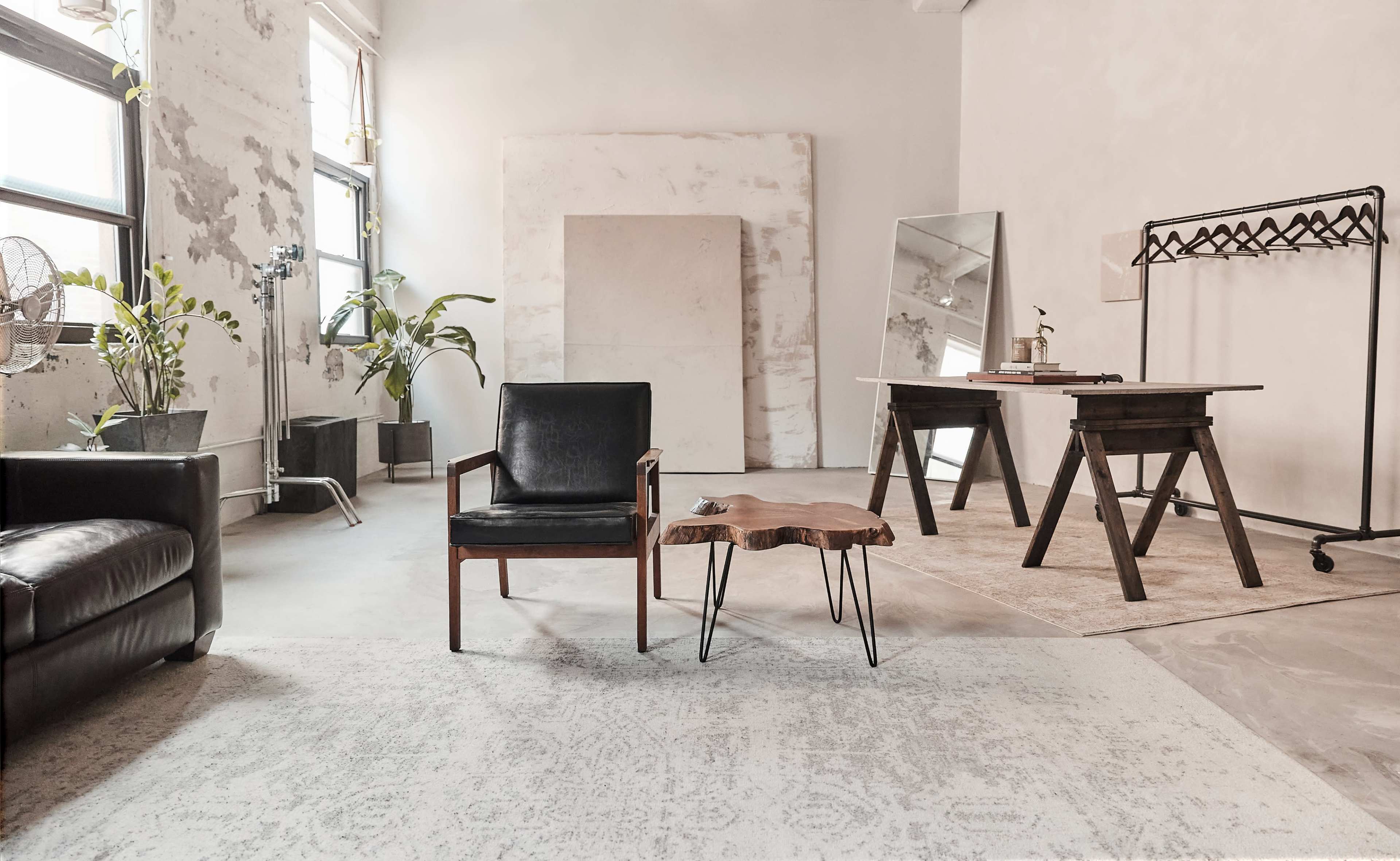 The image shows a modern, minimalist interior space featuring a black leather chair, a small wooden side table, a mirror, an industrial-style desk, and potted plants.