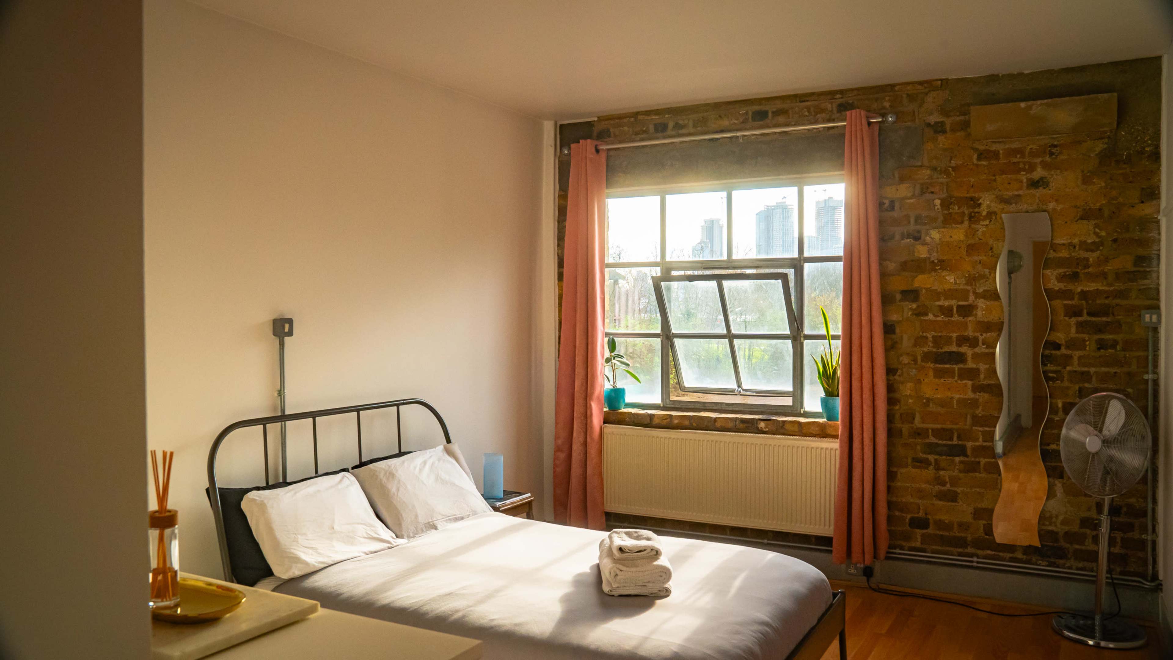 A well-lit bedroom with a bed, a window overlooking a cityscape, and a brick wall featuring a fan and potted plants.