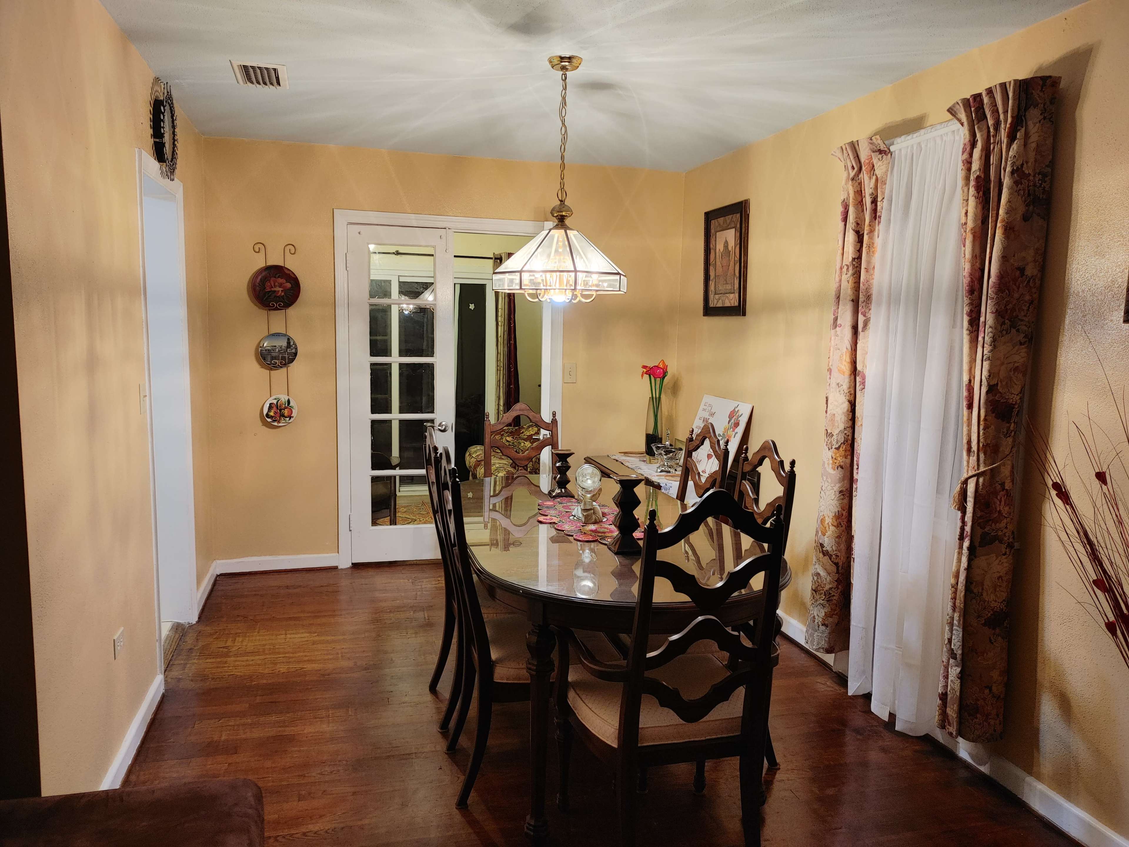 A dining room features a circular glass table surrounded by wooden chairs, illuminated by a pendant light, with a doorway leading to another room.