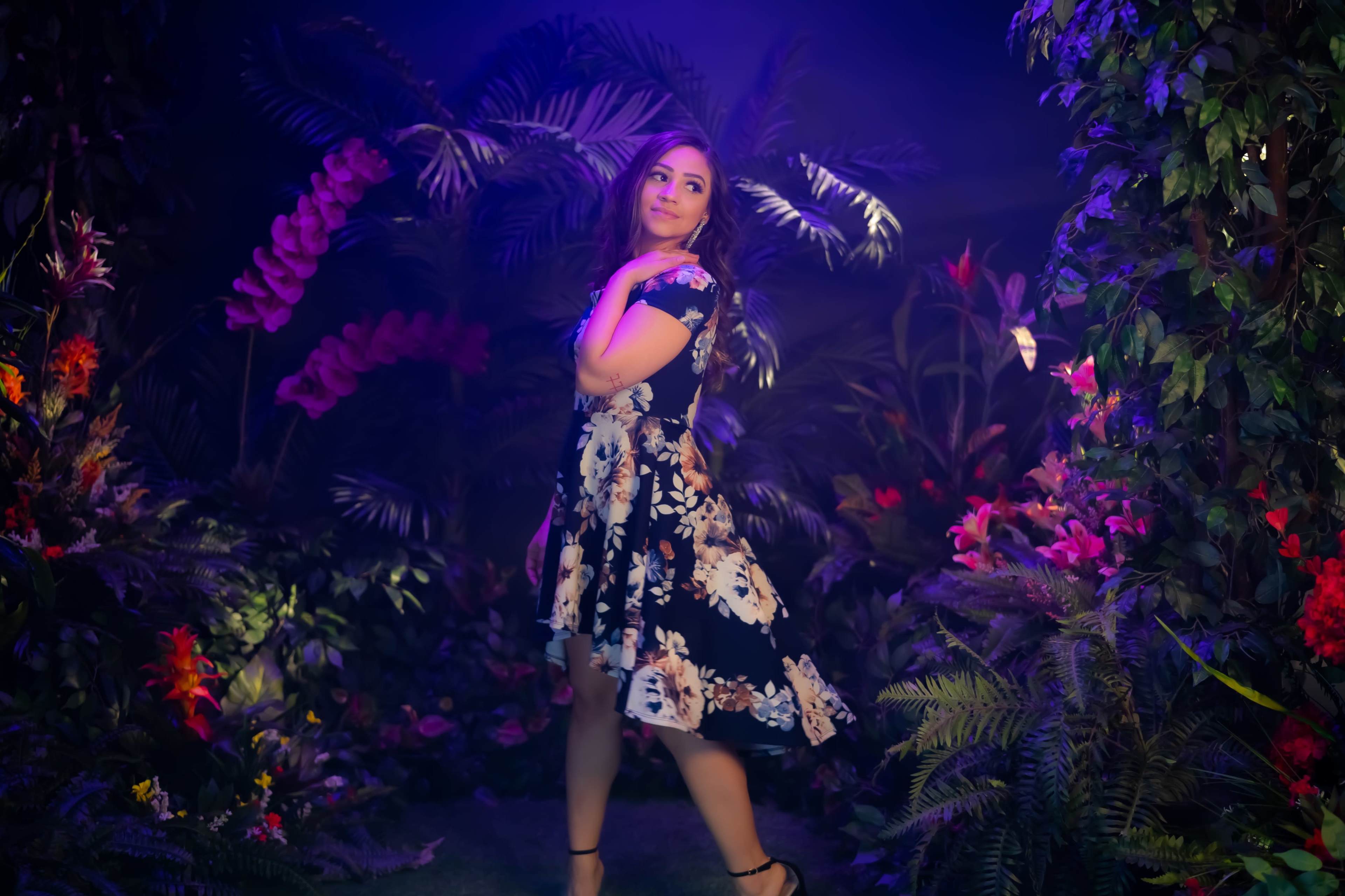 A woman poses elegantly in a floral dress within a vibrant, plant-filled environment illuminated by colorful lights.