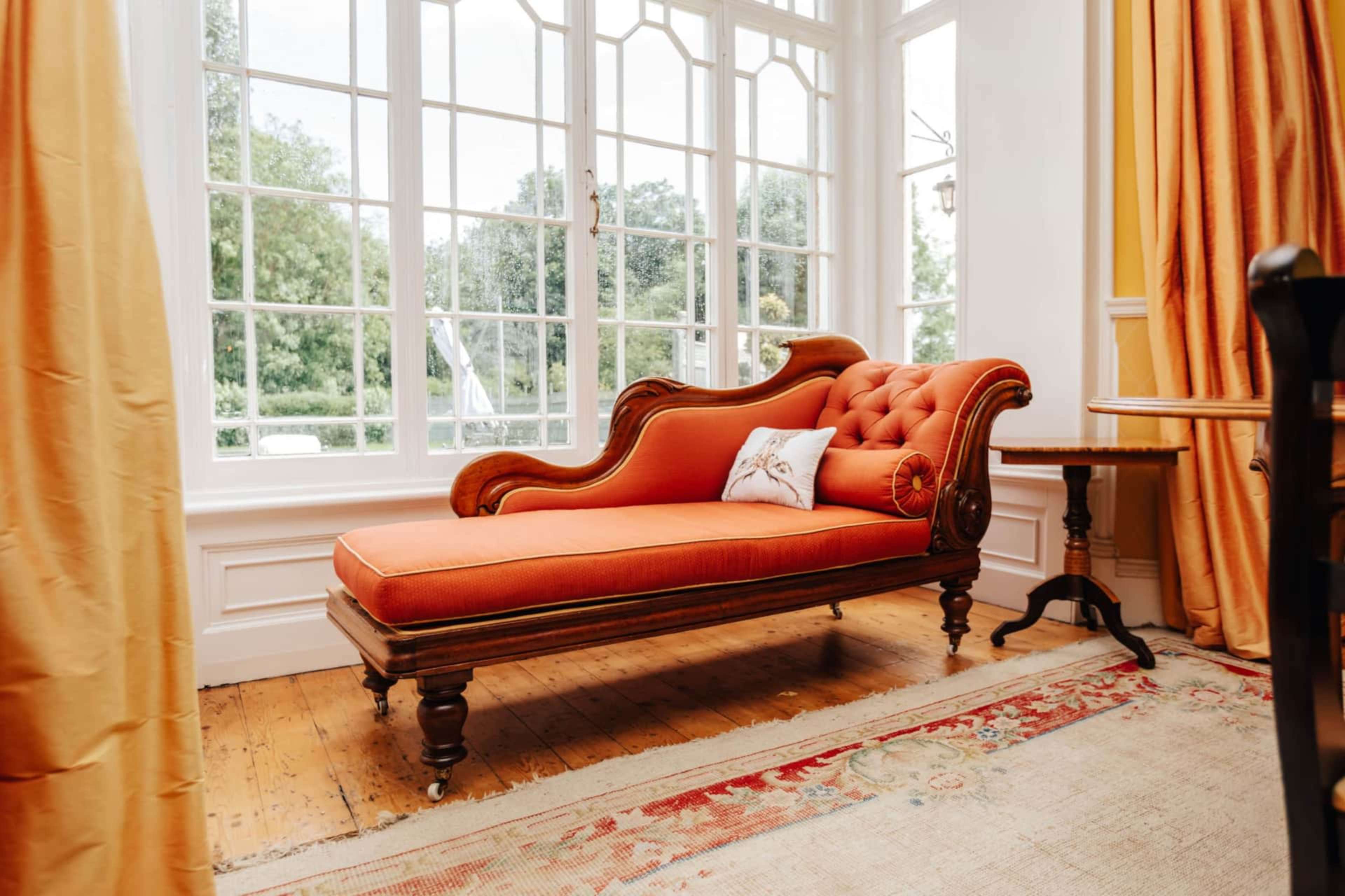 A vintage orange chaise lounge sits beside a wooden table in a bright room with large windows and draped curtains.