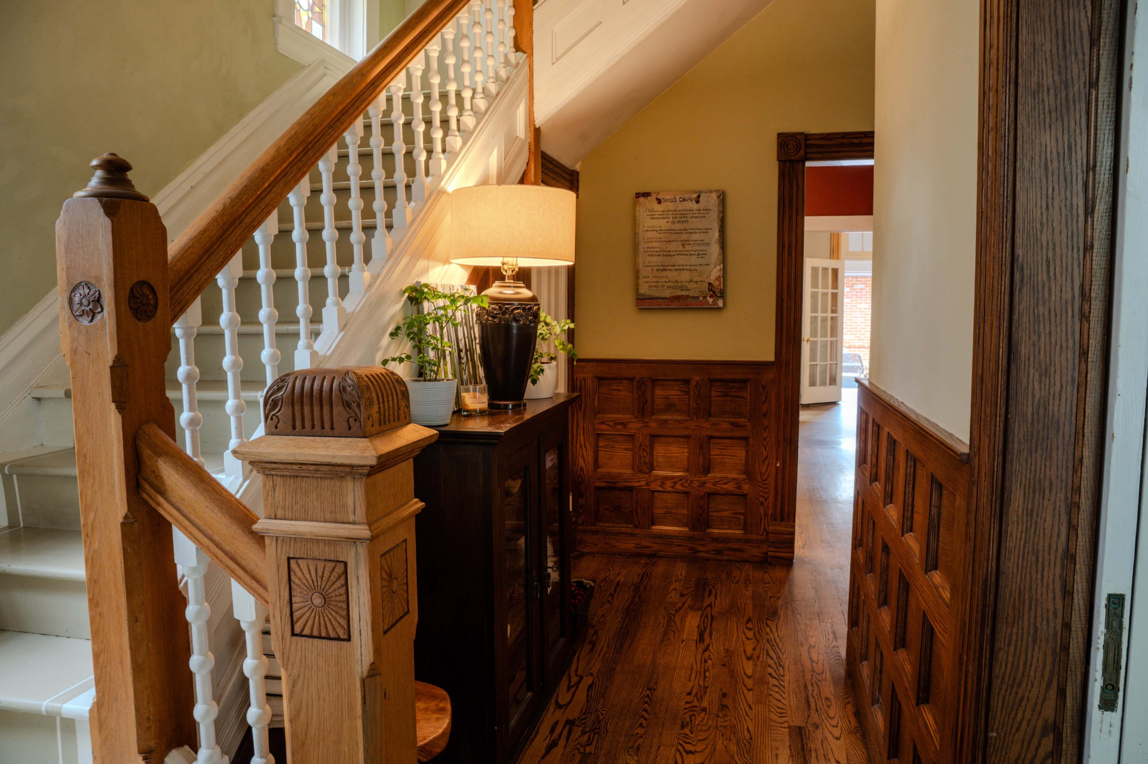 The image shows a well-lit hallway featuring a wooden staircase with a white banister, a console table with a lamp, and decorative wall paneling.