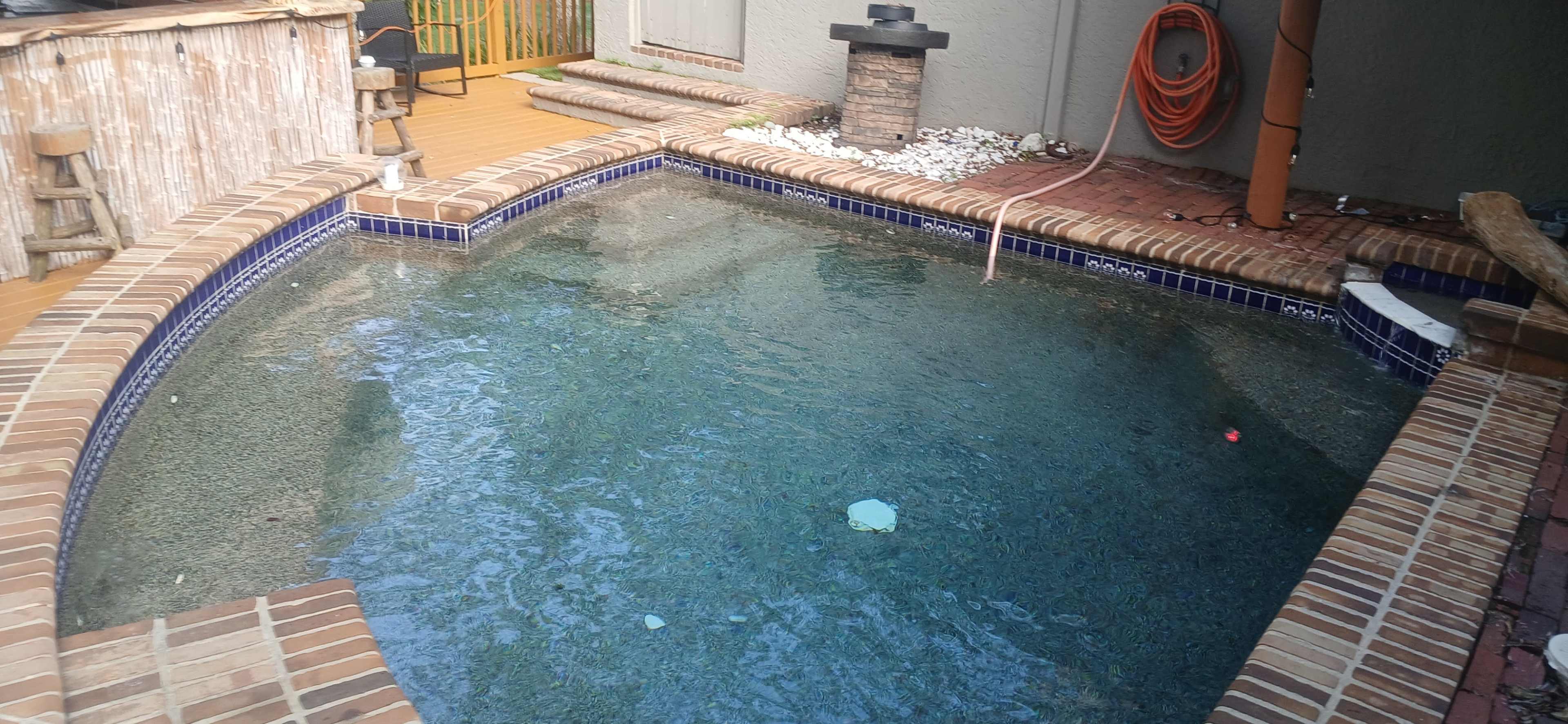 The image shows a rectangular swimming pool with a brick border, surrounded by outdoor features including a patio area and a stone fire pit.