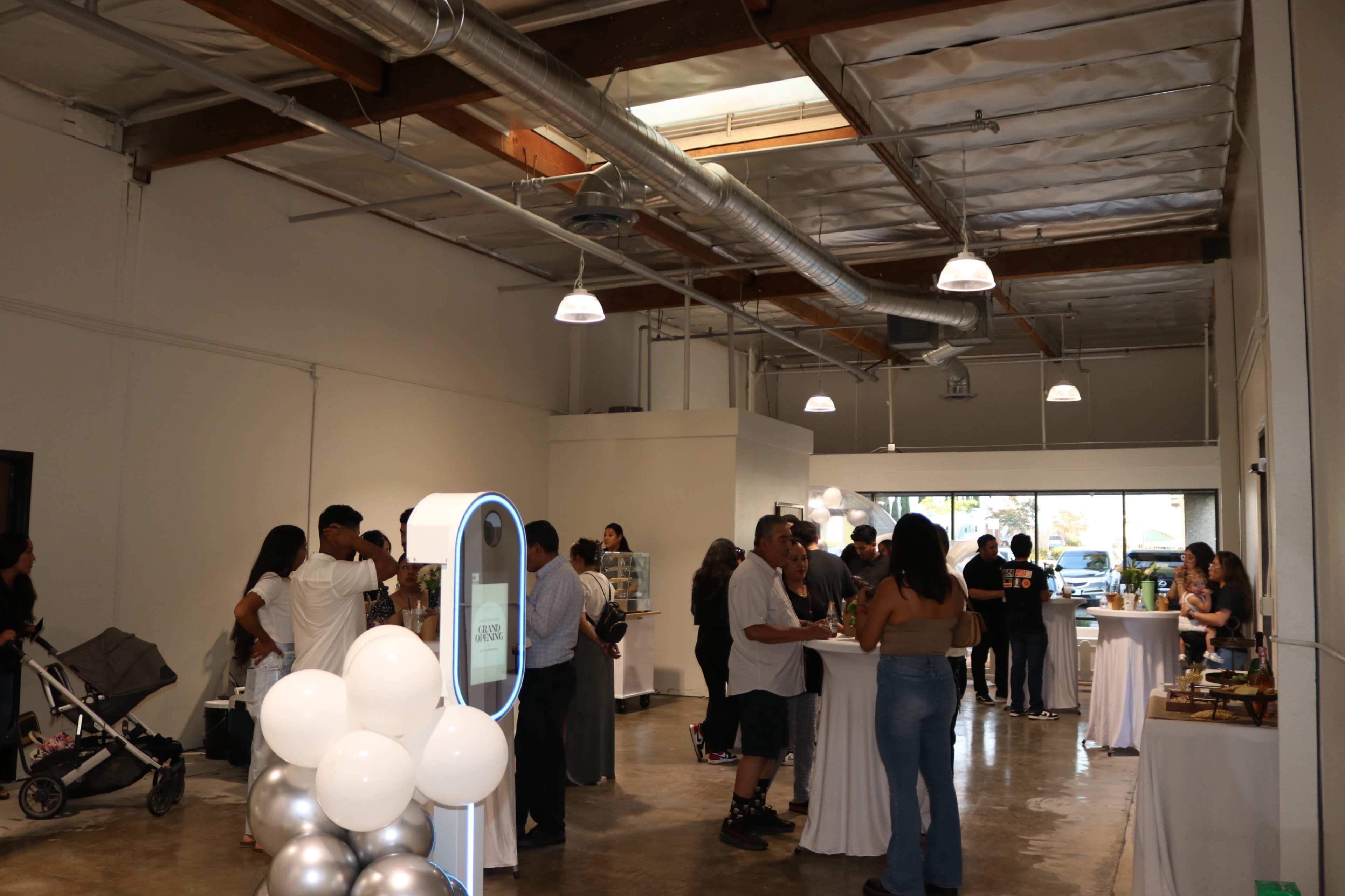 A group of people is gathered in a spacious, well-lit venue with high ceilings, mingling and enjoying refreshments at tables.