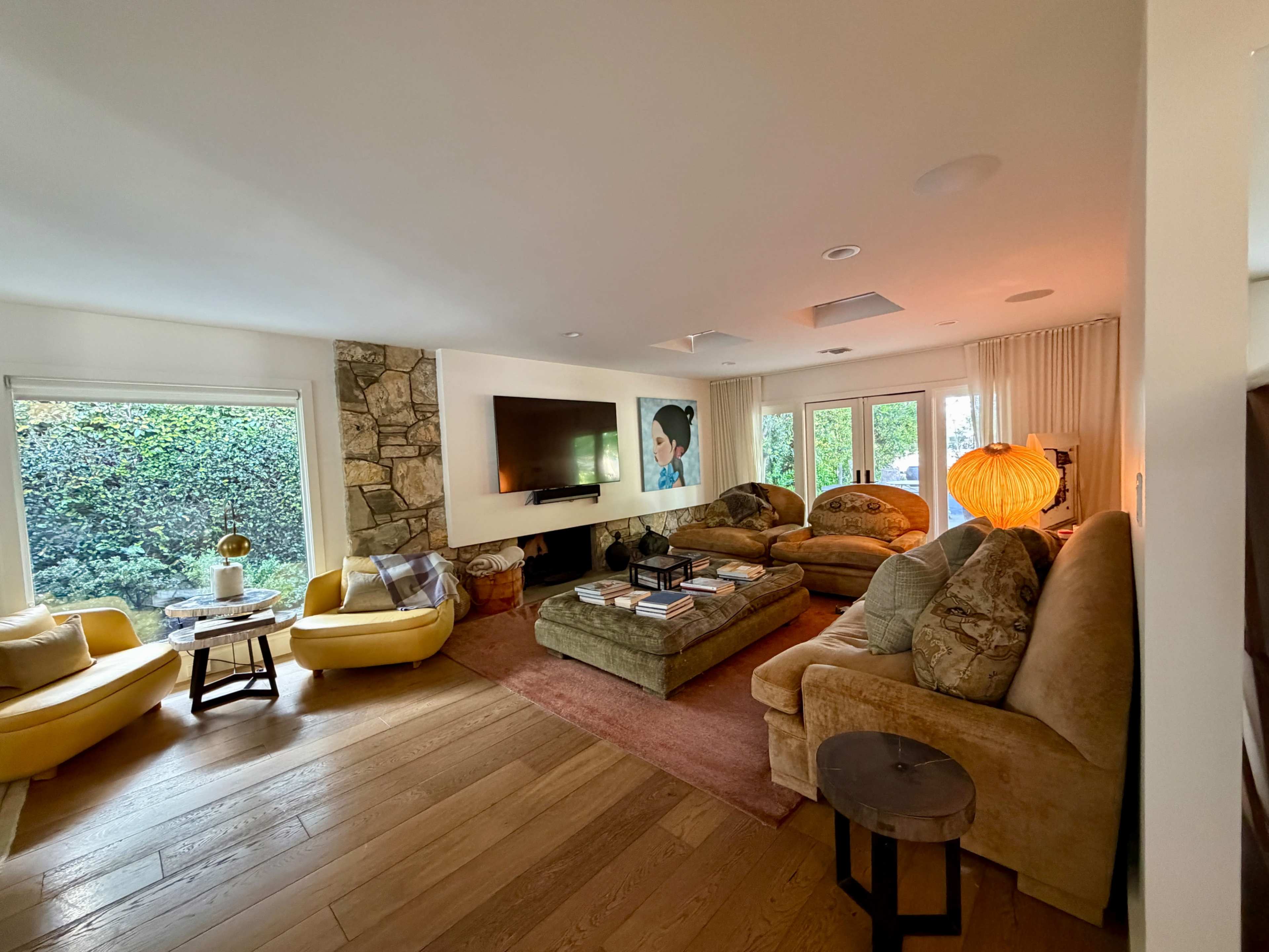 The image shows a spacious living room with multiple seating areas, a large television mounted on the wall, and windows revealing greenery outside.