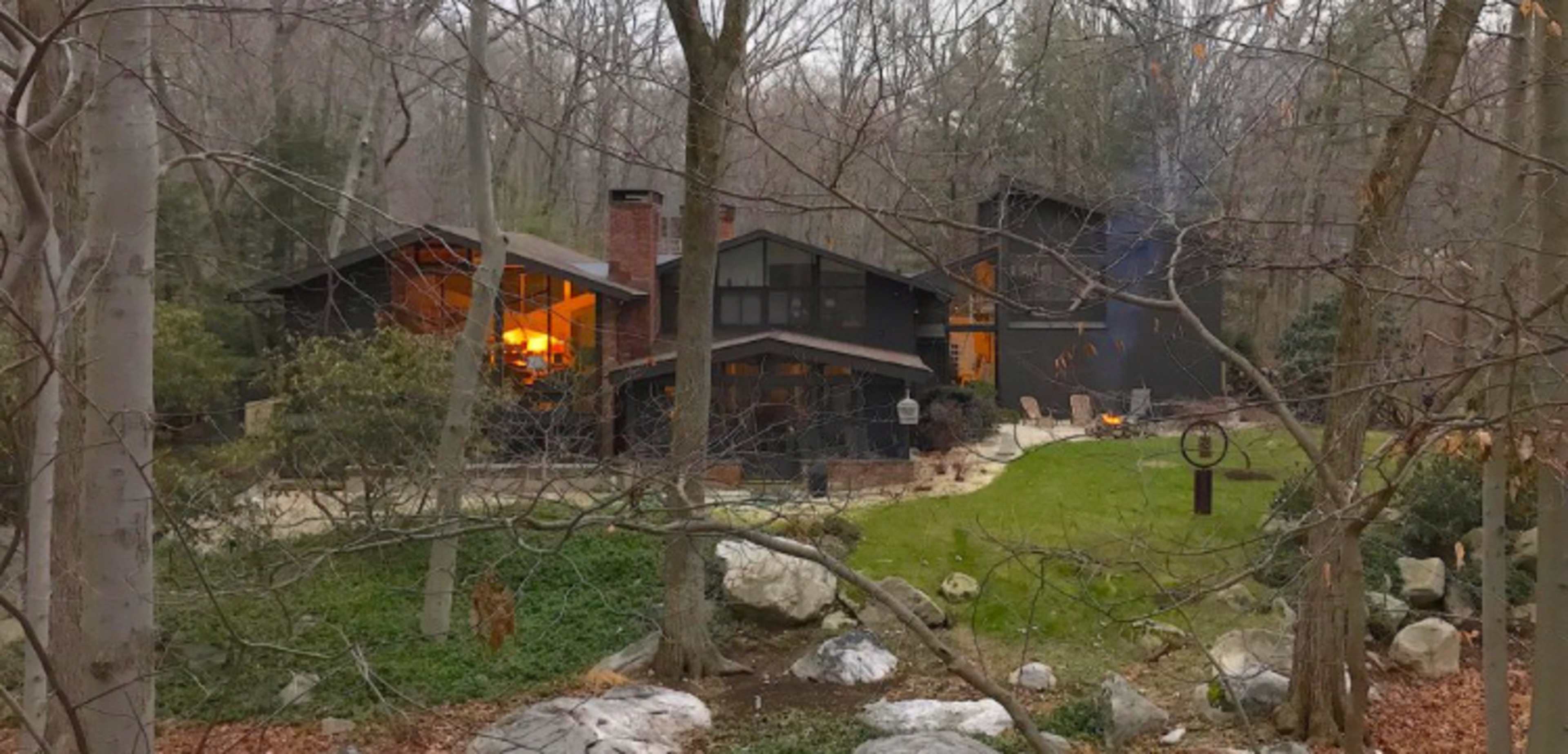 A modern black house surrounded by trees and rocks features warm light glowing from its windows.