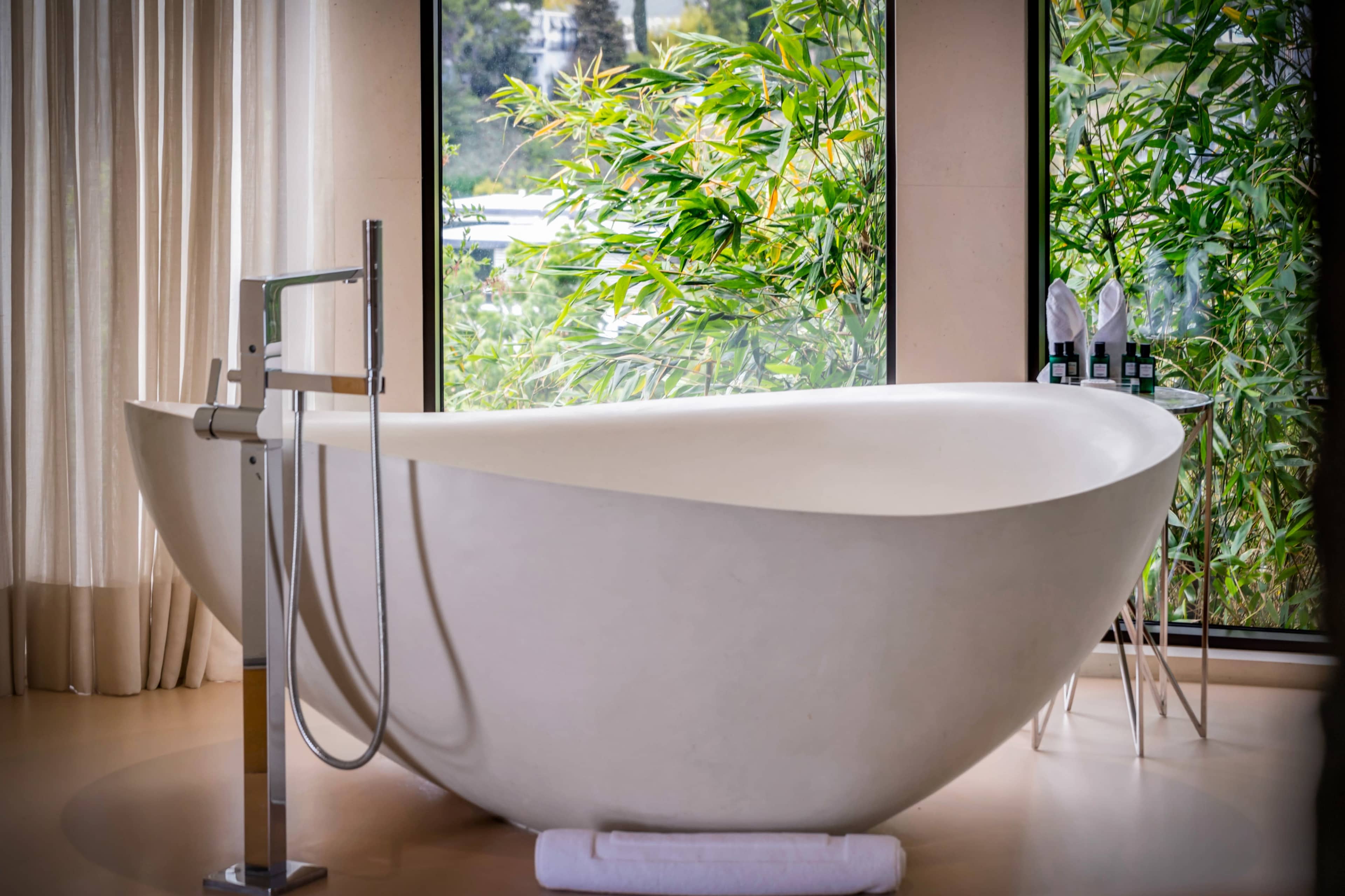 A sleek, modern freestanding bathtub is positioned in front of large windows with greenery visible outside.