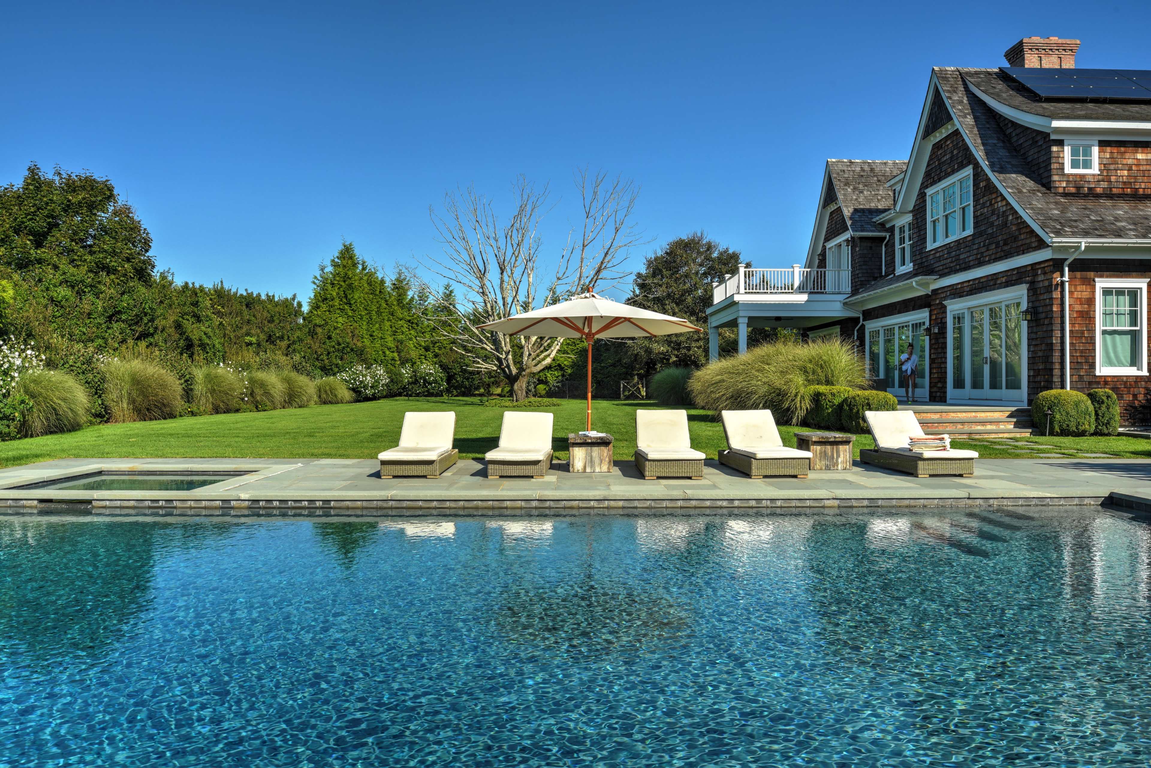 The image depicts a spacious backyard with a swimming pool, sun loungers, an umbrella, and a stylish house surrounded by greenery.