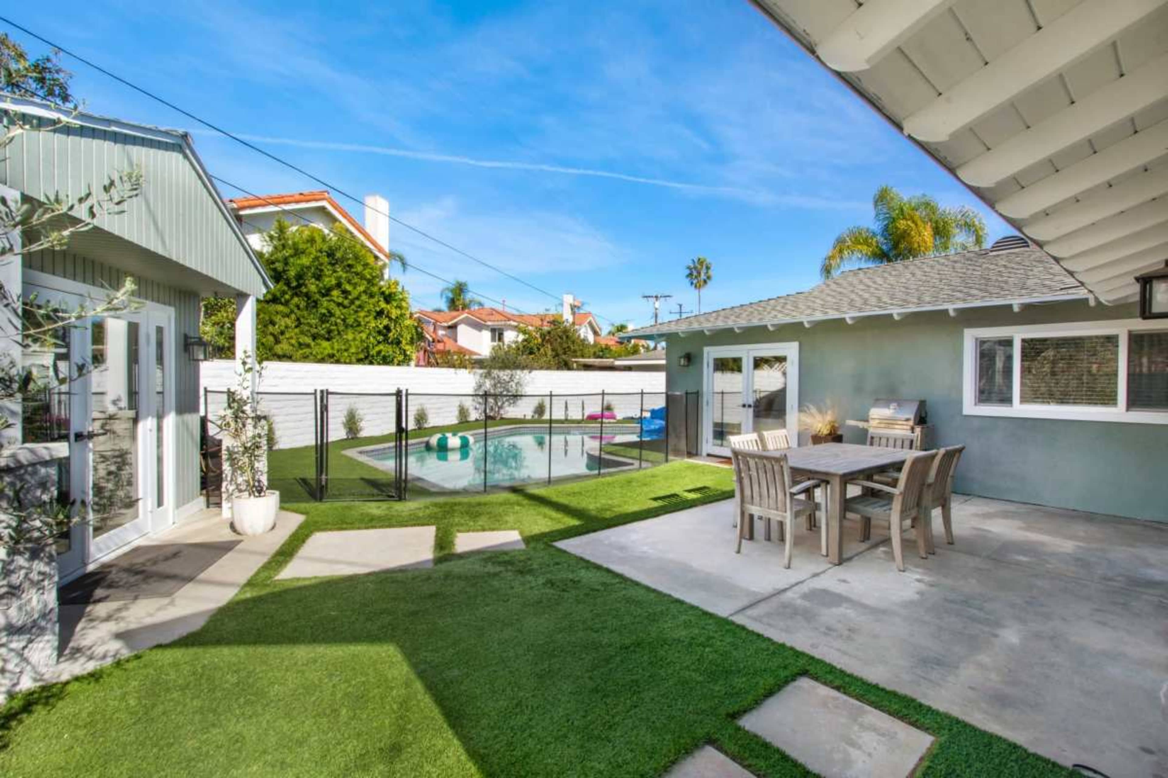 The image shows a backyard with a swimming pool surrounded by a fence, a seating area with table and chairs, and a small building in the background.