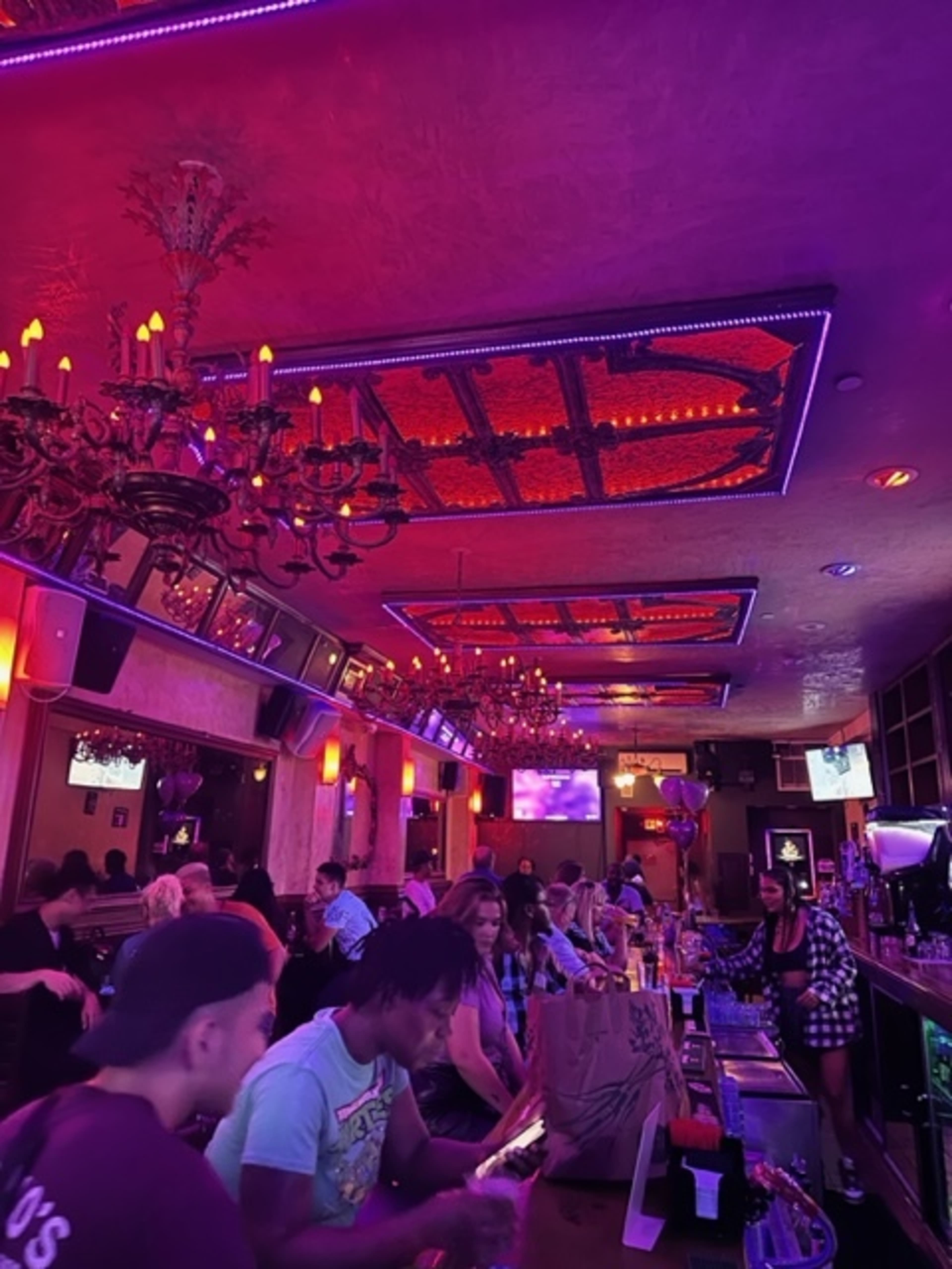A lively bar scene features patrons sitting at the counter and tables, illuminated by purple and red lights, with ornate chandeliers hanging from the ceiling.