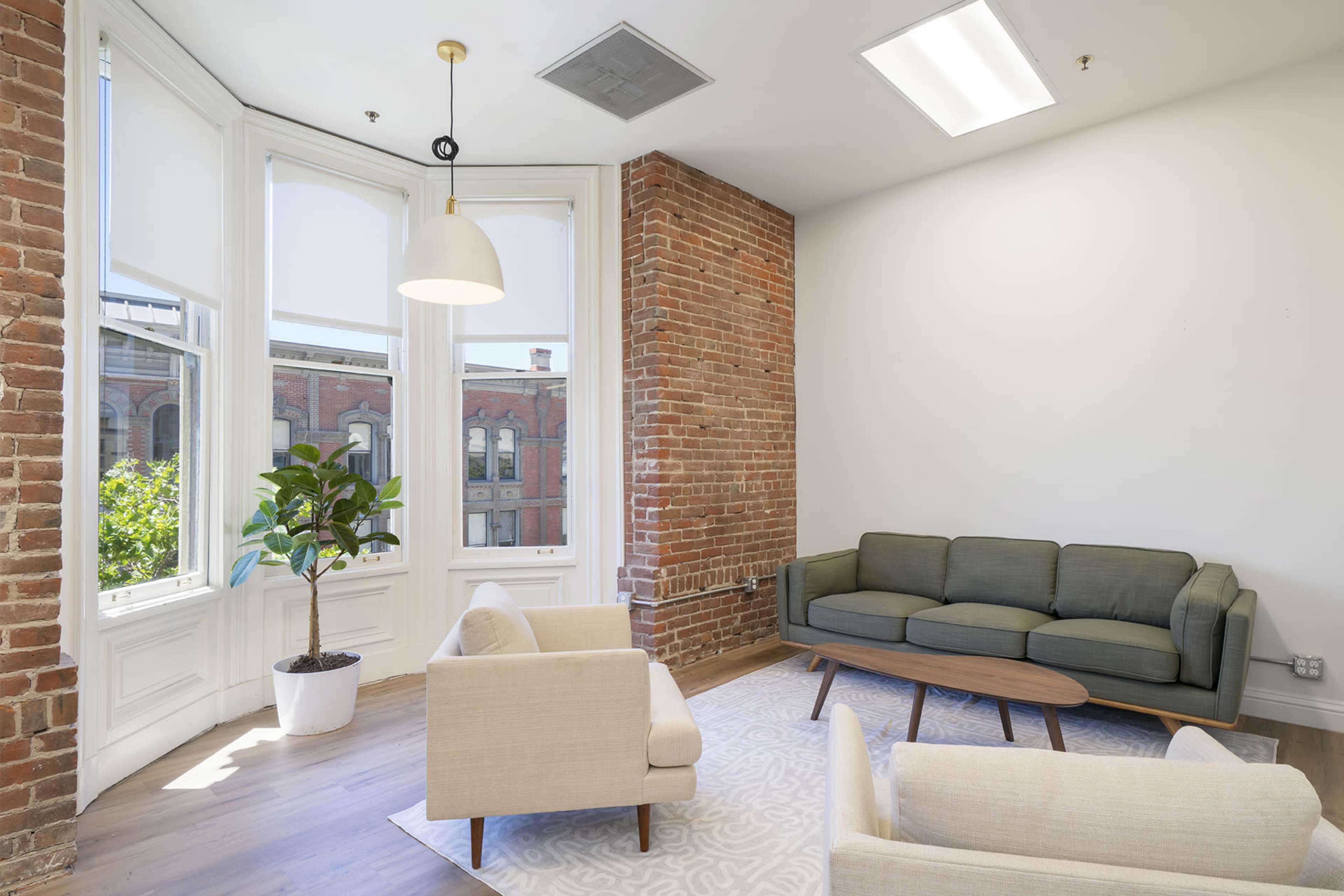 A cozy sitting area featuring a green sofa, two light-colored armchairs, a wooden coffee table, and a potted plant, with large windows letting in natural light and exposed brick walls.