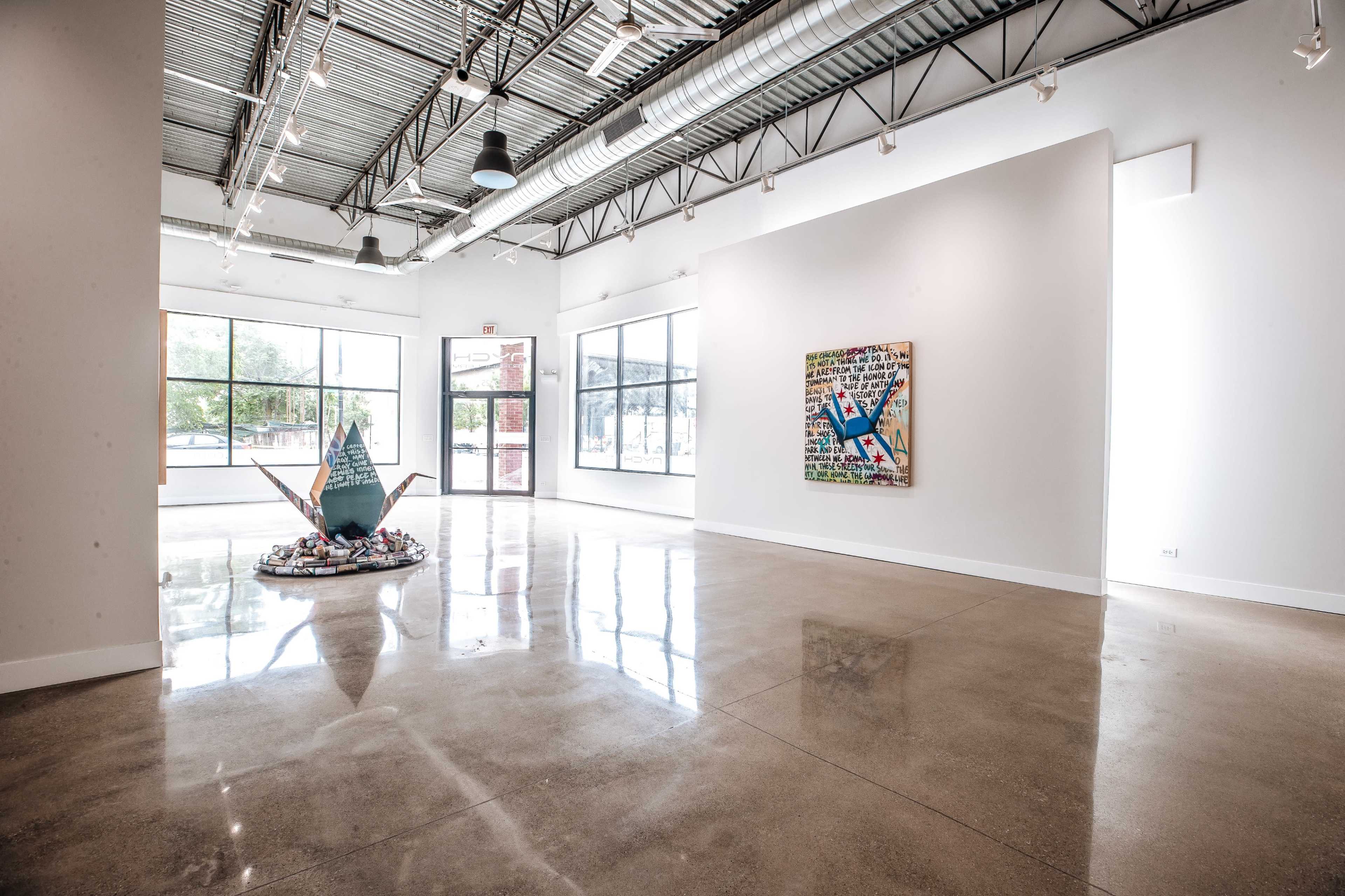 The image shows a spacious art gallery with large windows, polished concrete floors, and contemporary artworks displayed on the walls.