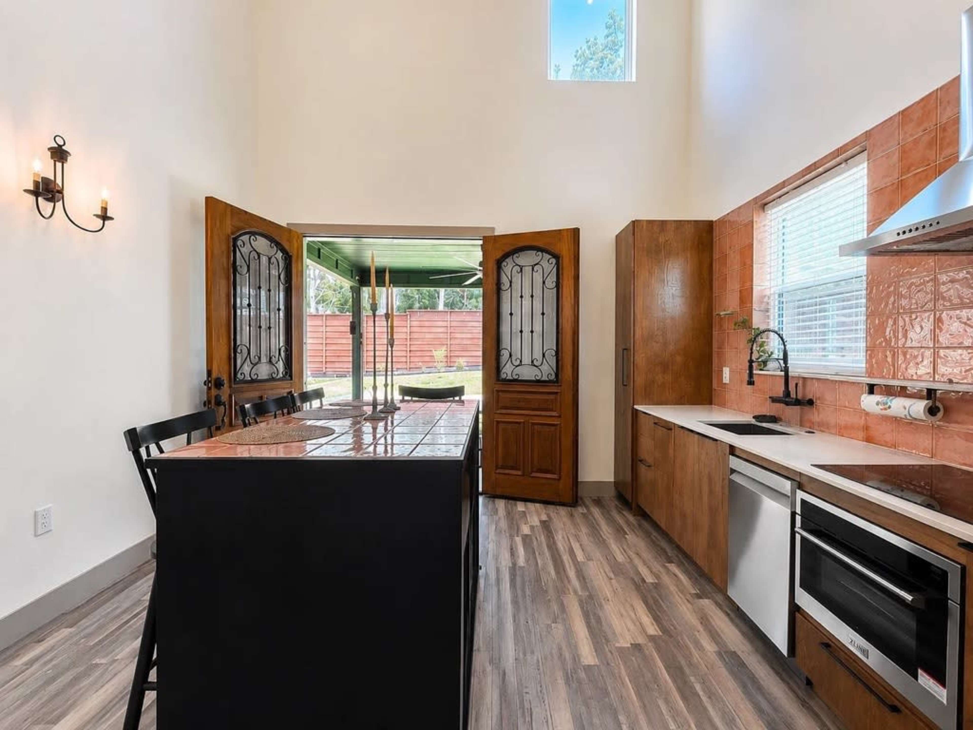 The kitchen features a central island with black countertops, wooden cabinetry, and large double doors leading to an outdoor area.