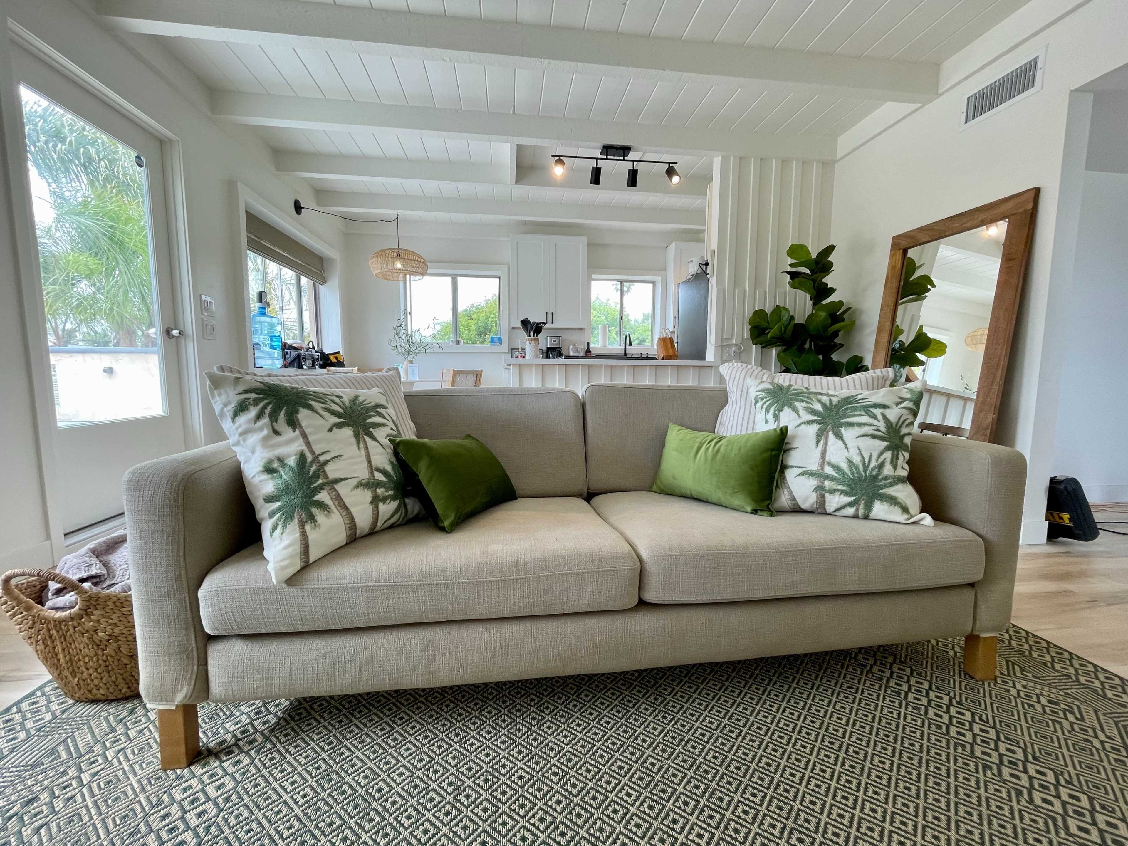A beige sofa with palm tree-patterned cushions sits on a patterned rug in a bright, open living space with large windows and a kitchen in the background.