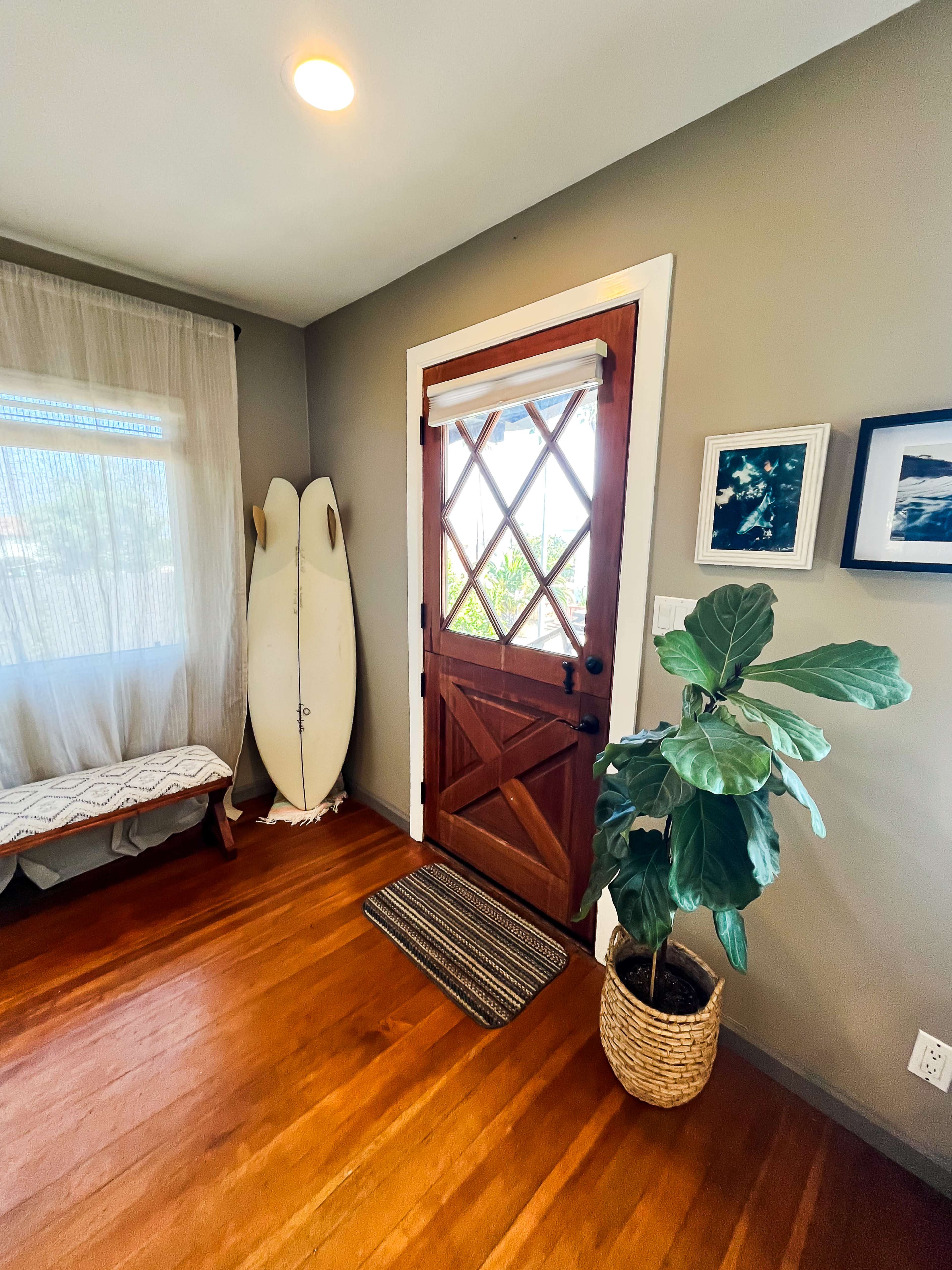 A welcoming entryway with a wooden door, a bench, a surfboard against the wall, and a potted plant.