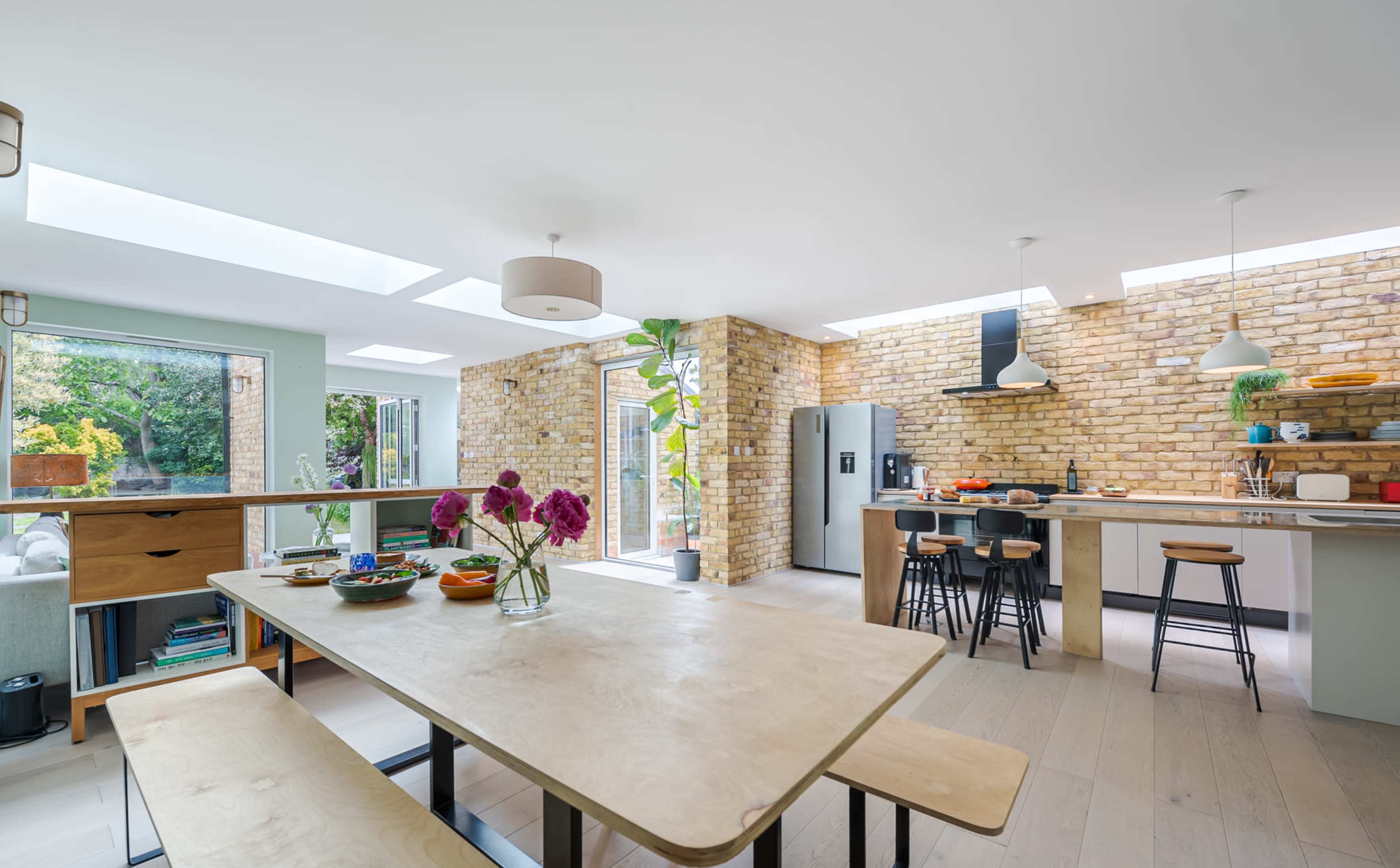 A modern kitchen and dining area featuring a wooden dining table, brick walls, and large windows letting in natural light.