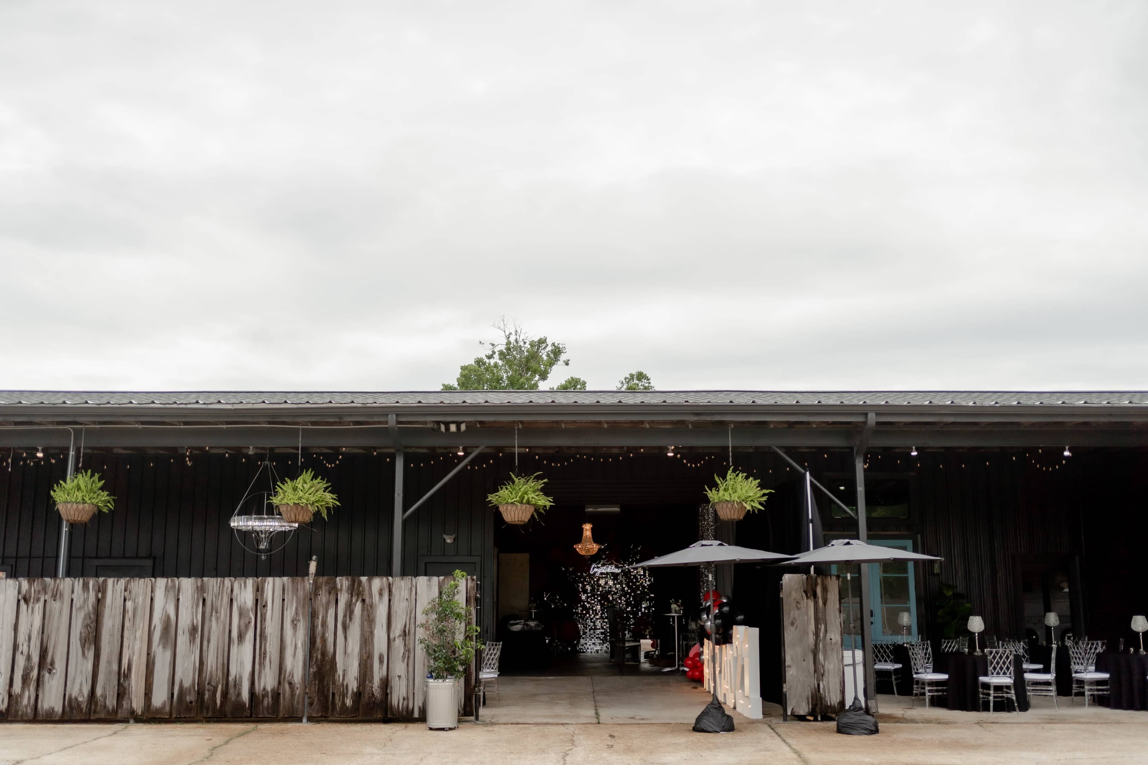 A rustic building with a dark exterior, featuring hanging plants, outdoor seating with umbrellas, and festive lighting above an entrance.