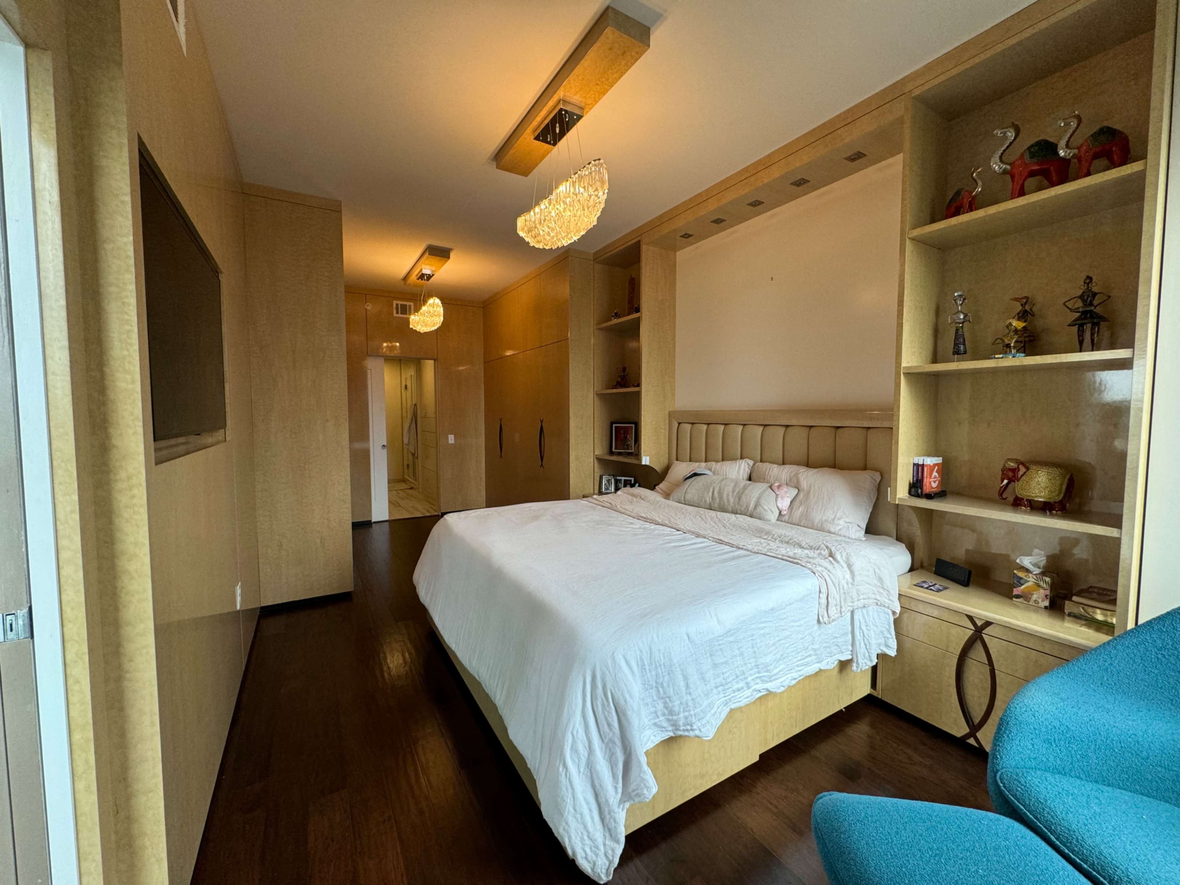 The image shows a modern bedroom featuring a bed with a light-colored wooden headboard, a built-in shelving unit, and a small blue chair in a corner.