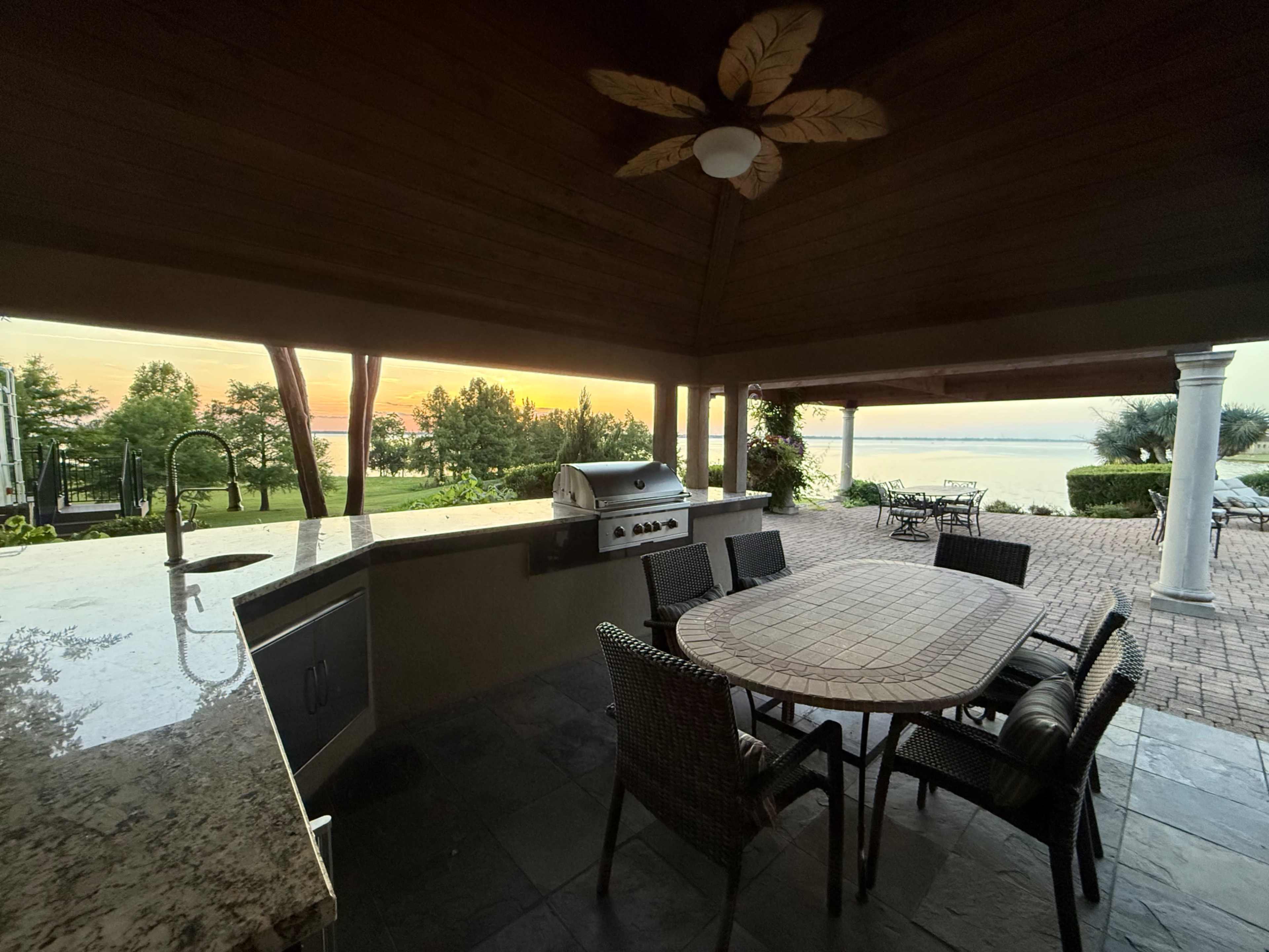 An outdoor kitchen under a covered patio overlooks a serene waterfront at sunset.