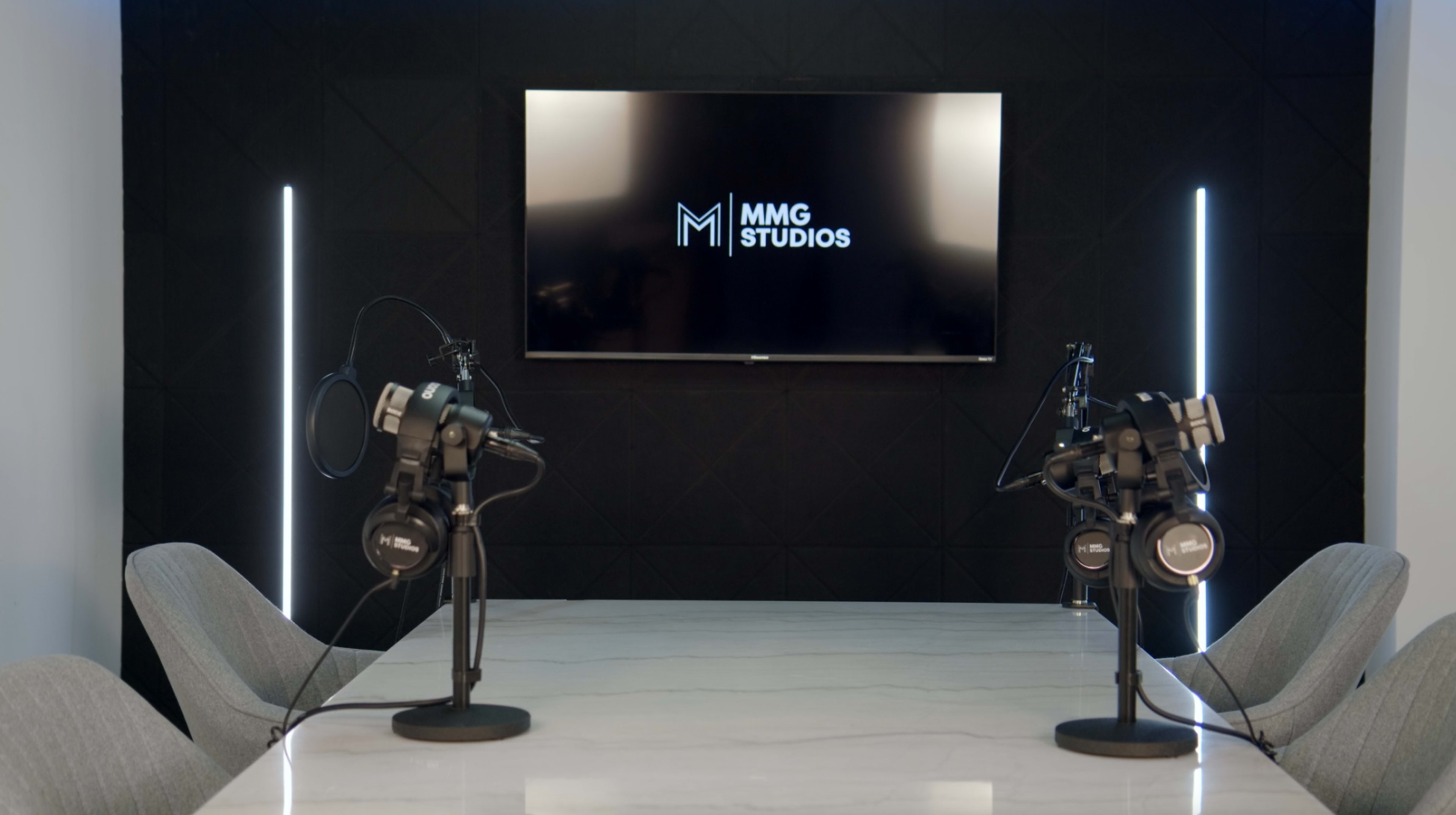 A podcast recording studio with two microphones and headphones positioned on a marble table, facing a screen displaying the "MMG Studios" logo.