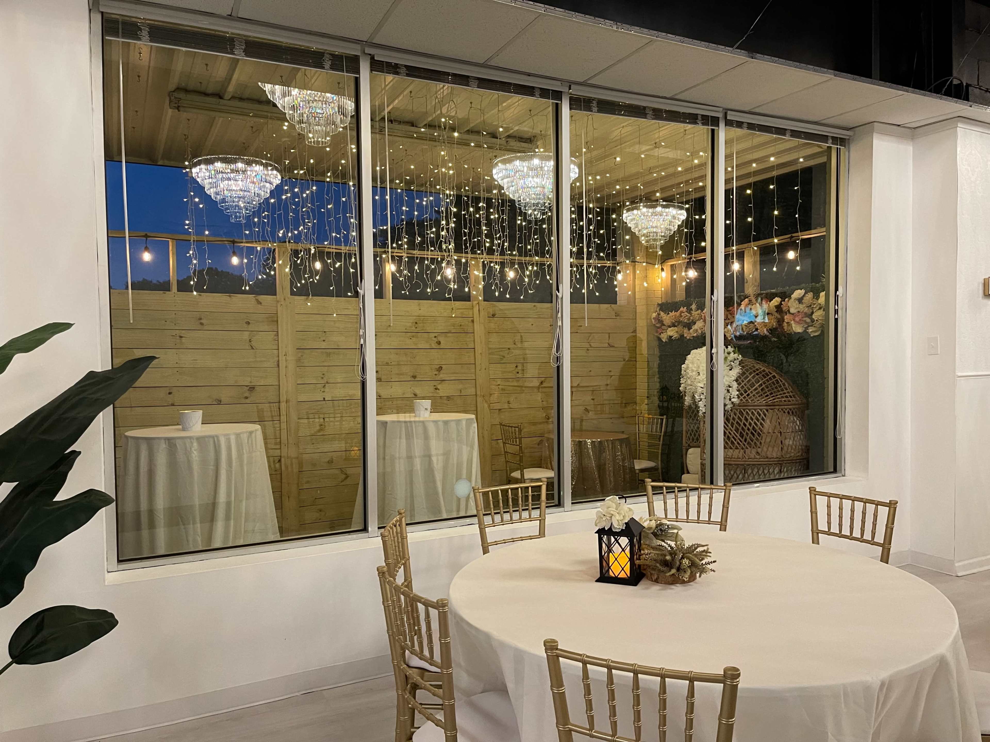 A spacious room with two elegant chandeliers and twinkling string lights, featuring round tables covered with cream tablecloths, viewed through large windows.