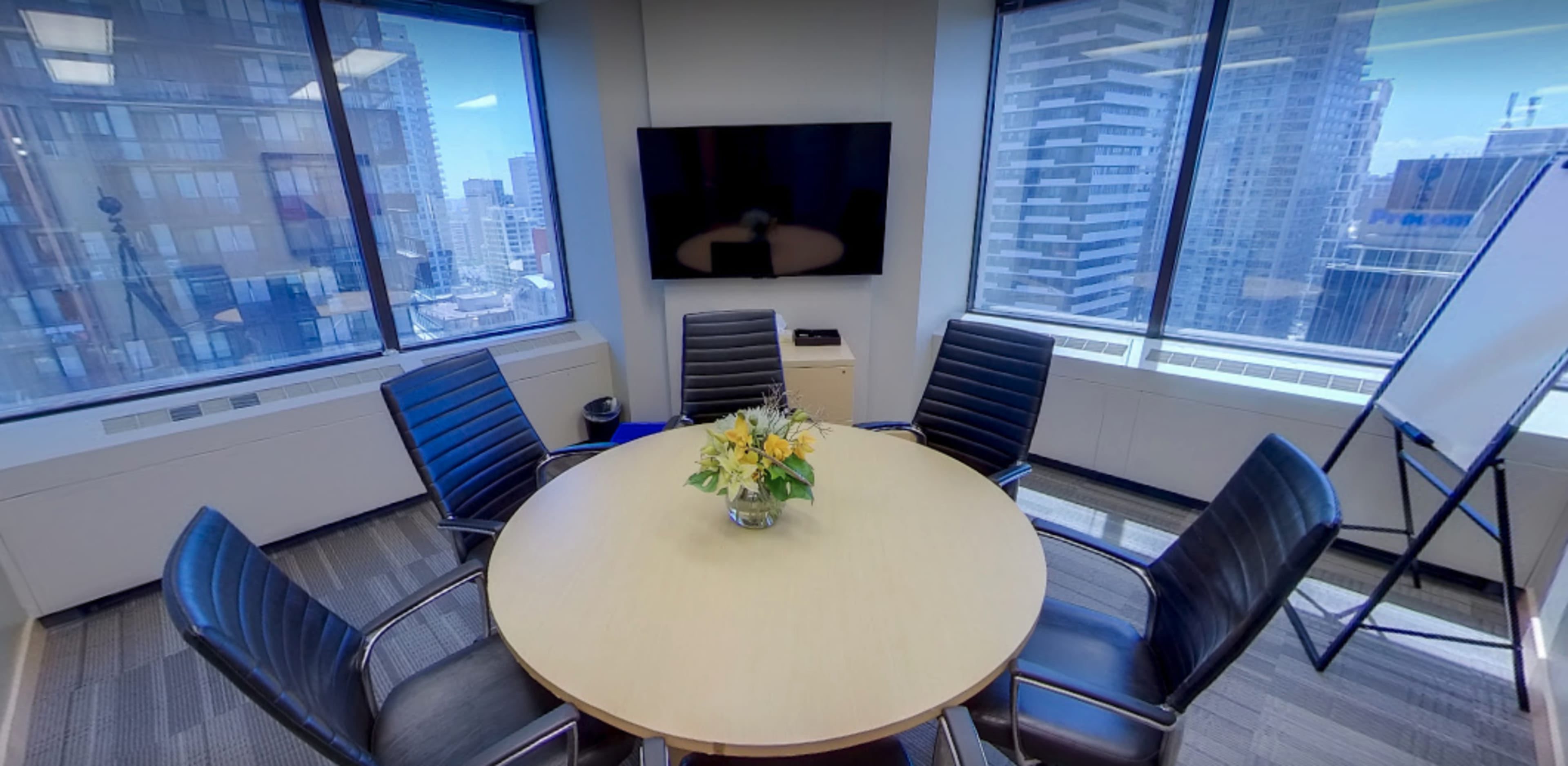 A conference room features a circular table surrounded by black chairs, large windows providing city views, and a wall-mounted television.