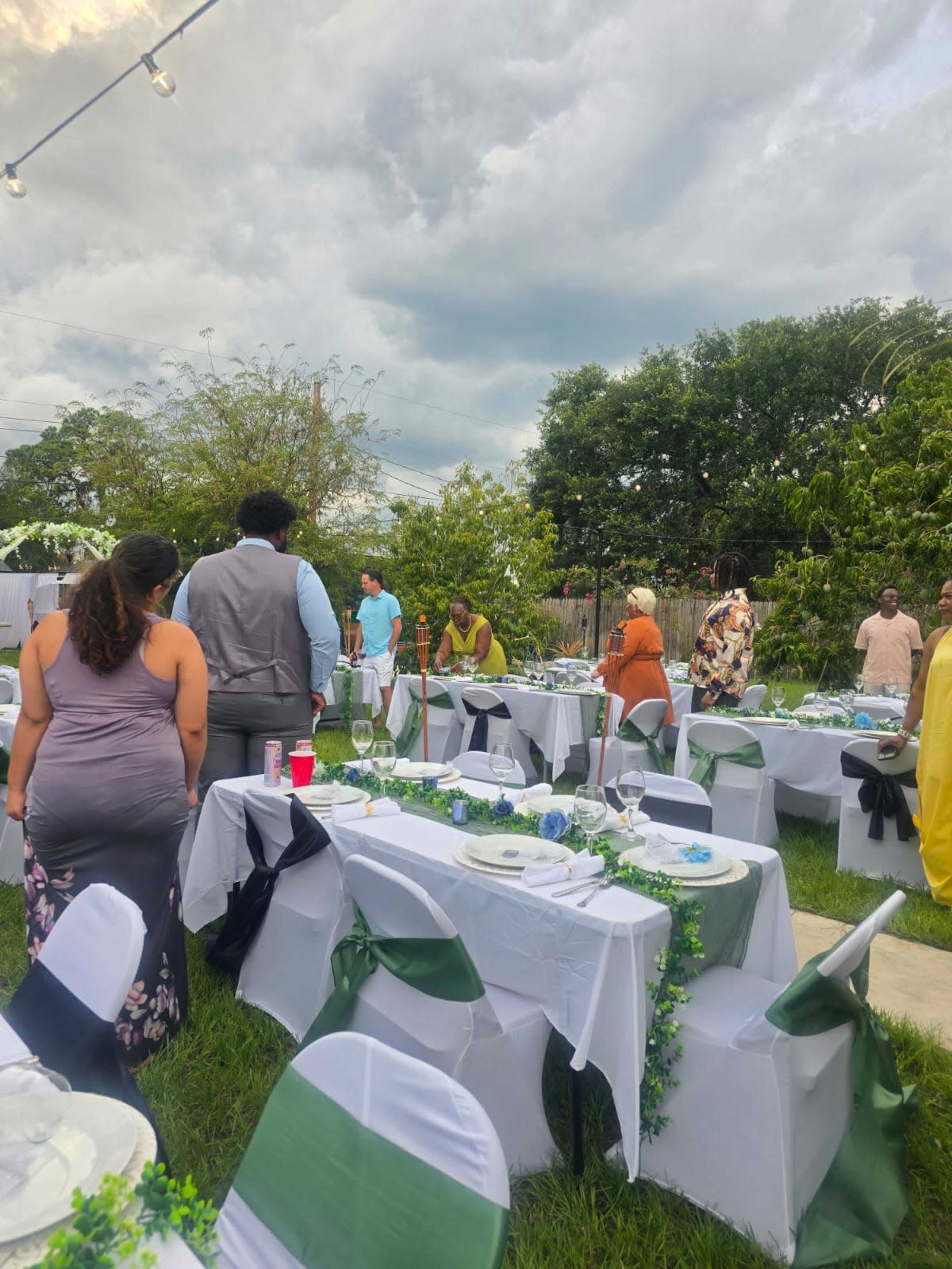 Guests gather around elegantly set tables in a backyard decorated for an outdoor event, with greenery and string lights overhead.