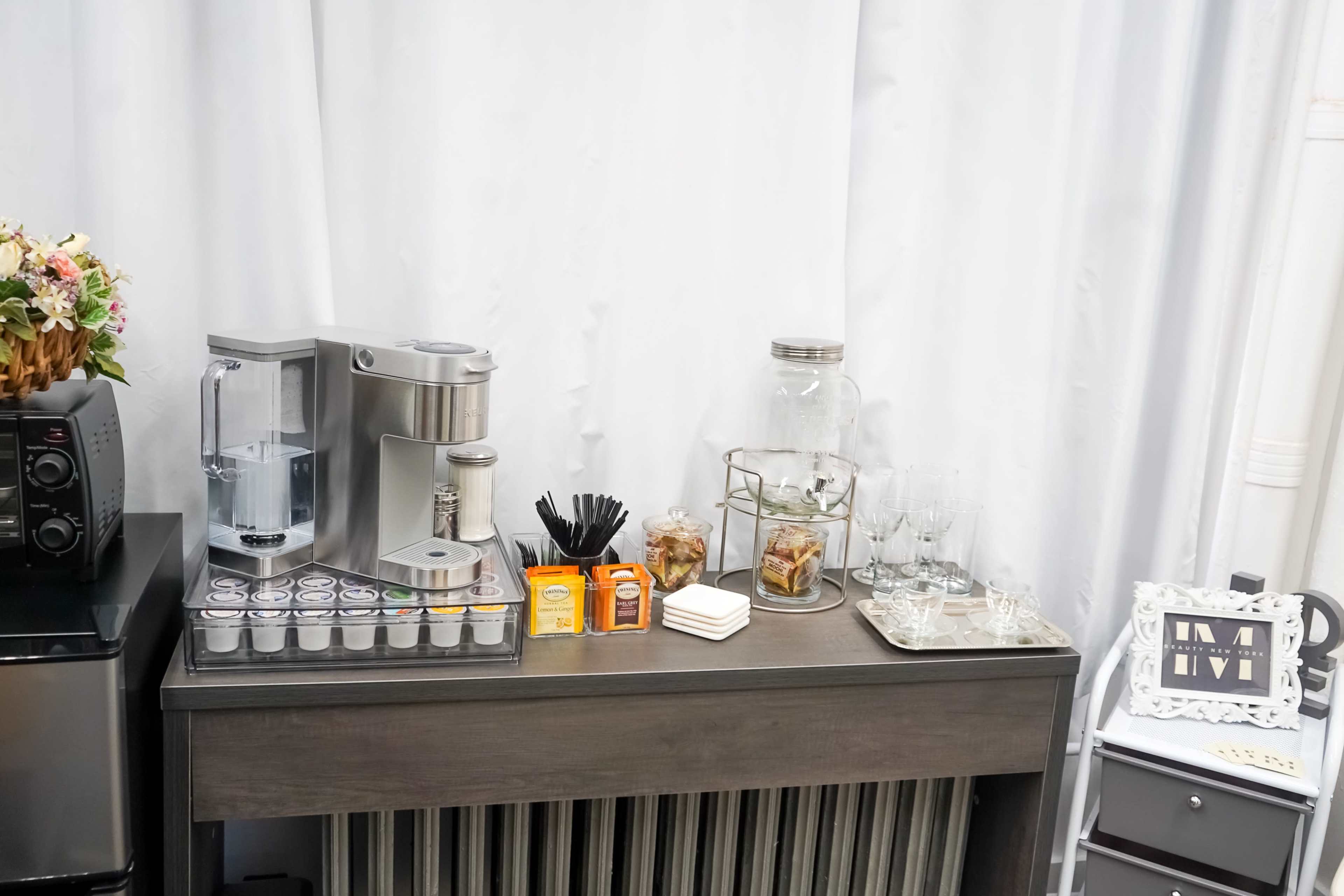 A beverage station features a coffee machine, a selection of tea and coffee pods, glass jars with snacks, utensils, and drinking glasses on a gray table against a white backdrop.